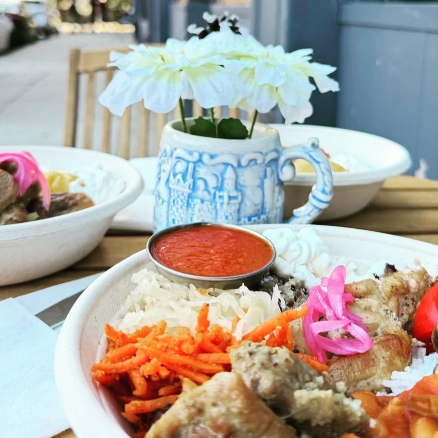 A colorful platter featuring rice, grilled meat, and a variety of vegetables, accompanied by a small bowl of red sauce, set on a table with a flower vase.