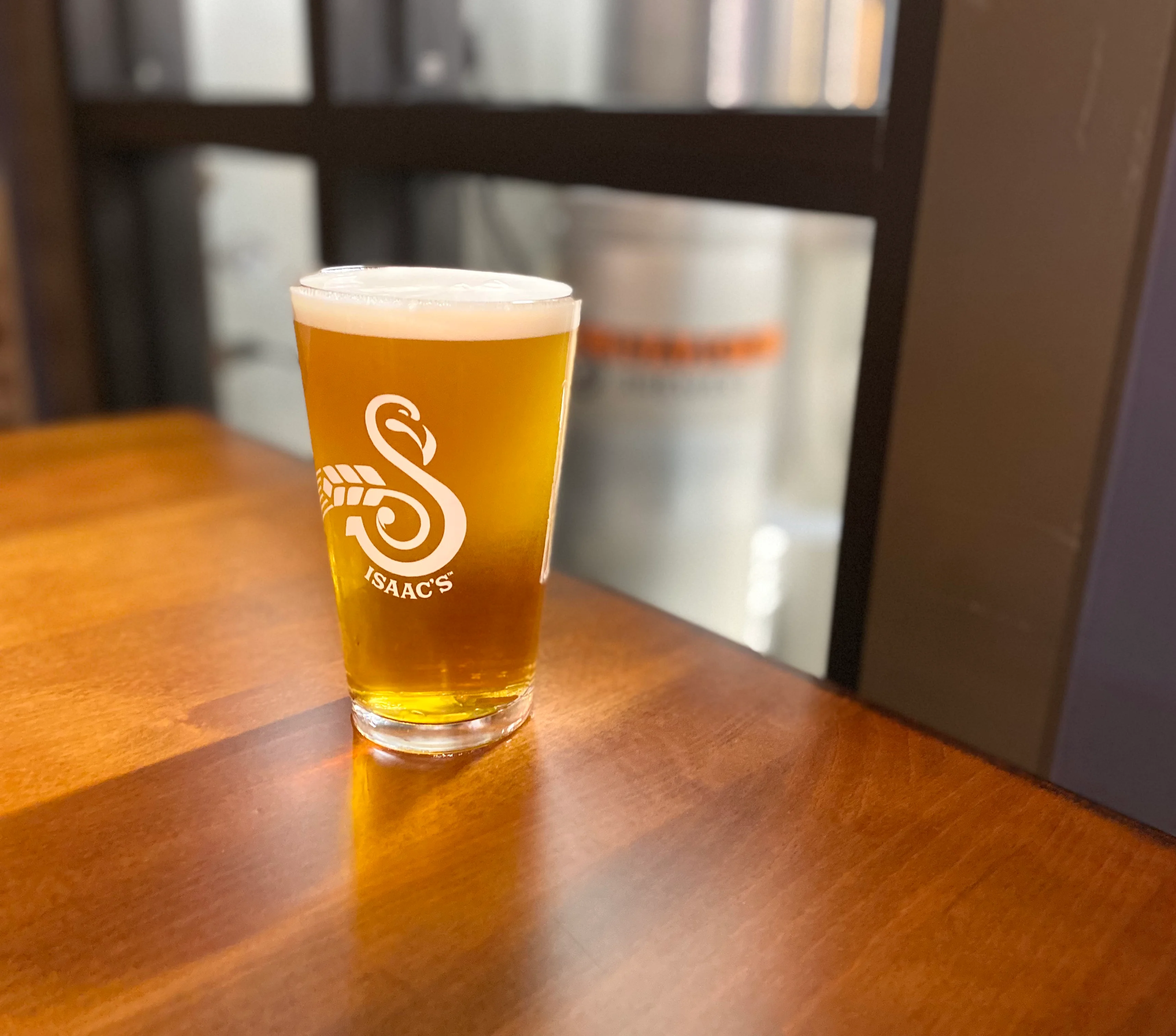 A pint of craft beer at Isaac's Craft Kitchen & Brewery, served in a branded glass with a golden amber hue and frothy head, set against the backdrop of the brewery's stainless steel tanks.
