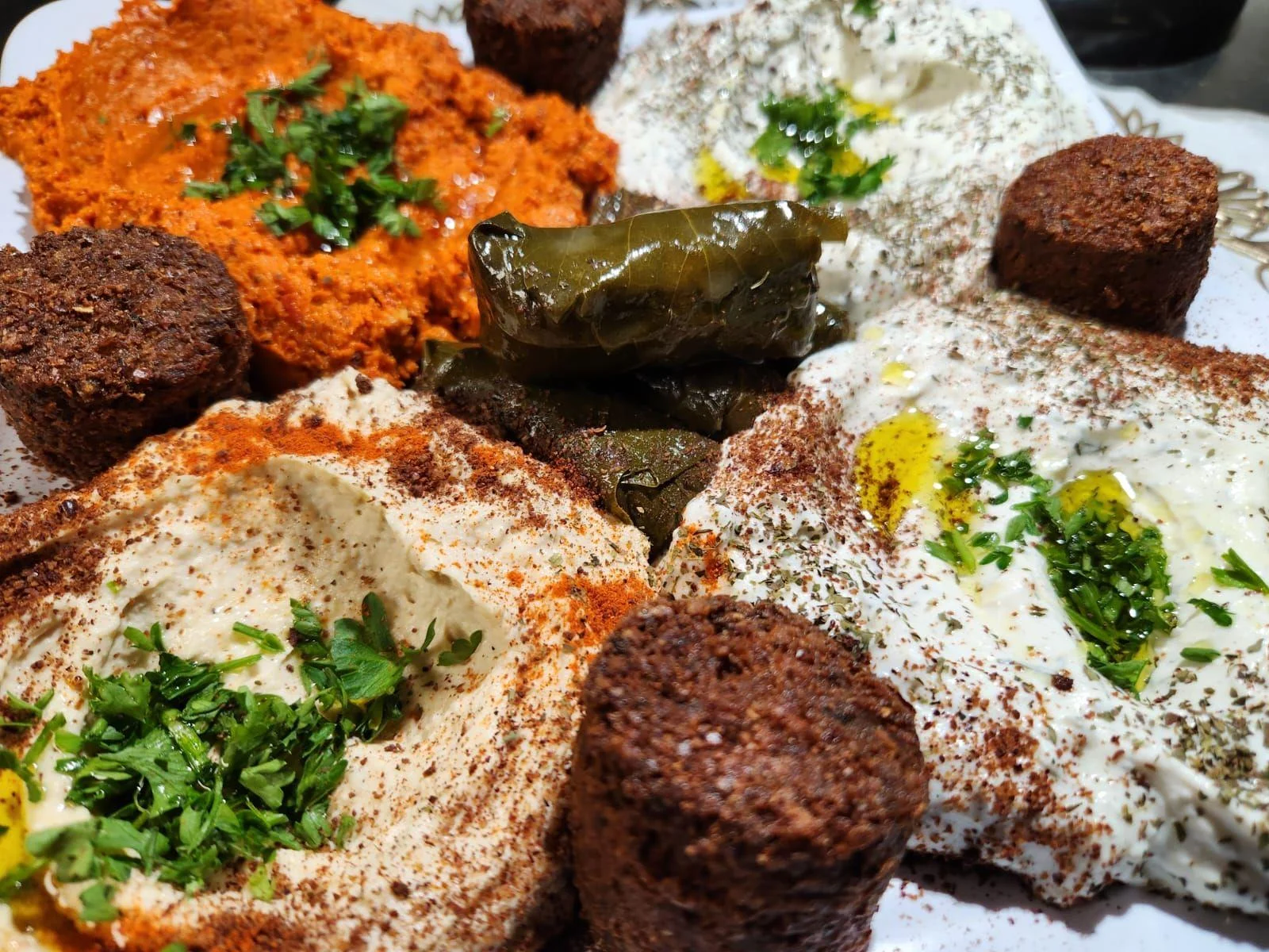 A platter featuring various dips, including hummus and spicy red spread, accompanied by grape leaves and small fritters.