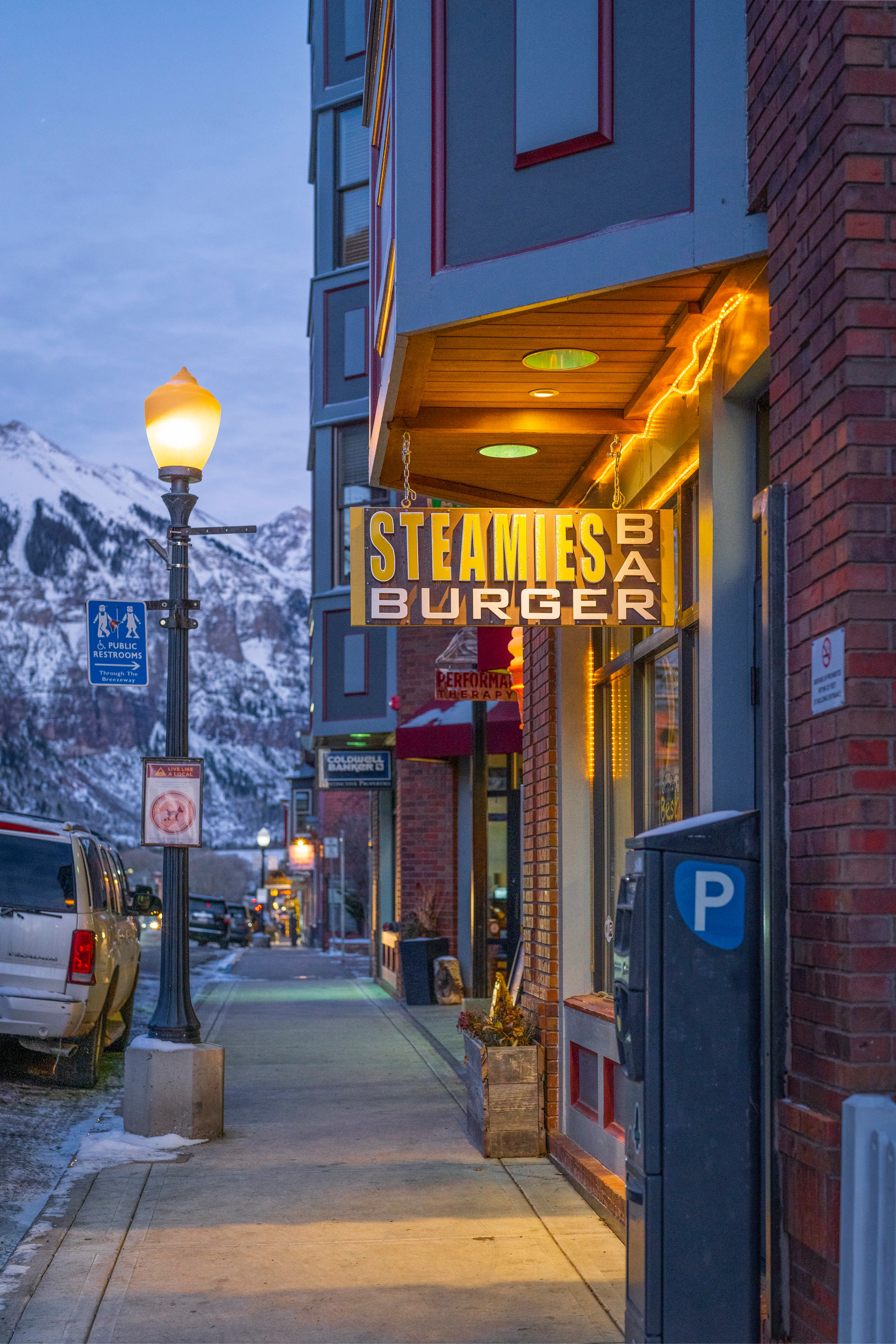The exterior of a burger restaurant with a sign illuminated in the evening light, set against a mountain backdrop.