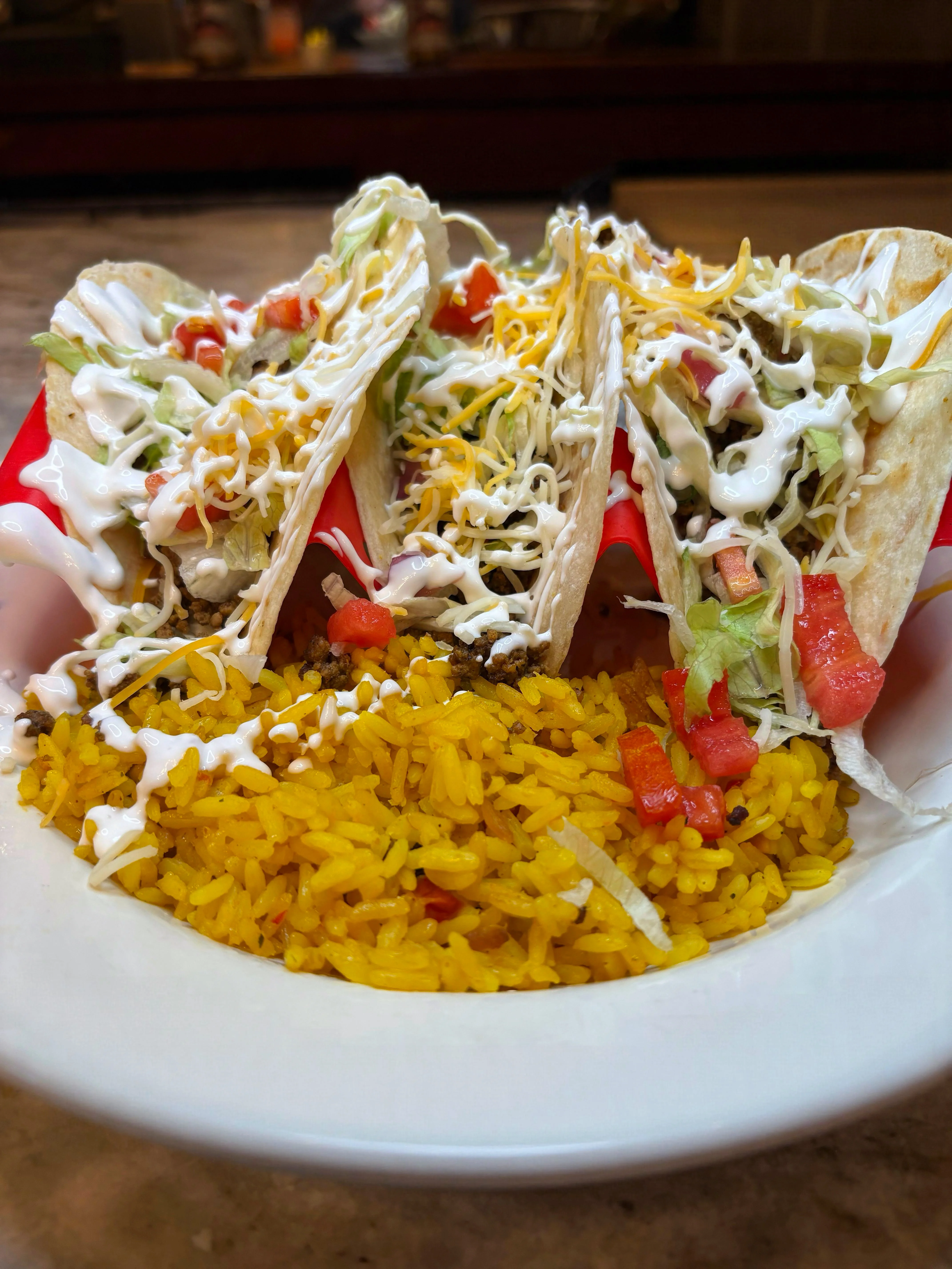 Three tacos filled with meat, lettuce, diced tomatoes, cheese, and topped with sour cream, served alongside a portion of seasoned yellow rice.