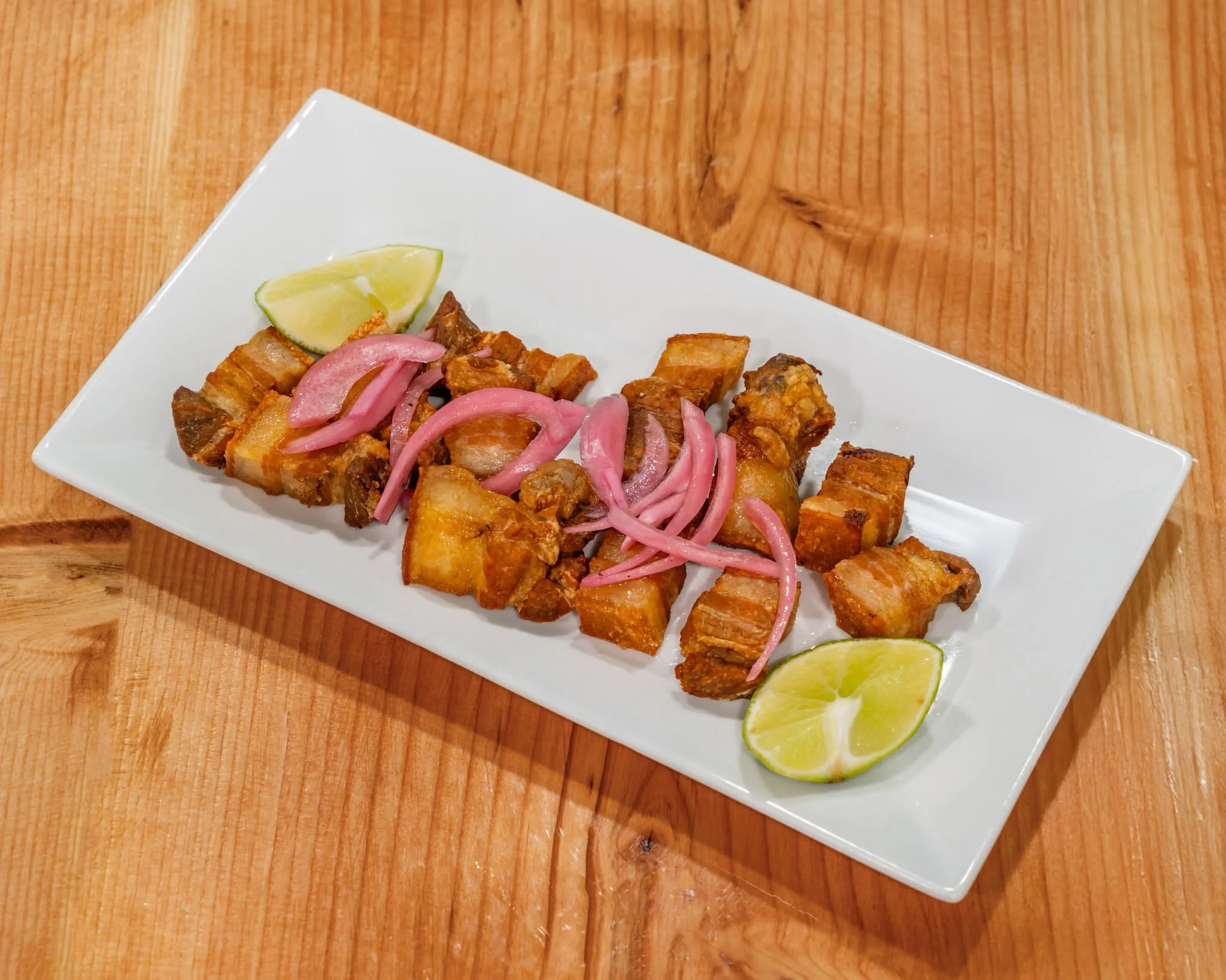A rectangular white plate of chicharrones, or fried pork belly, garnished with pickled red onions and lime wedges, resting on a wooden surface.