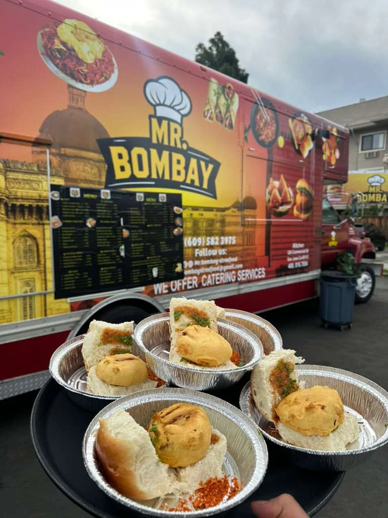 A food truck offering a variety of Indian dishes, showcasing small bread rolls filled with a spiced potato mixture on a tray.