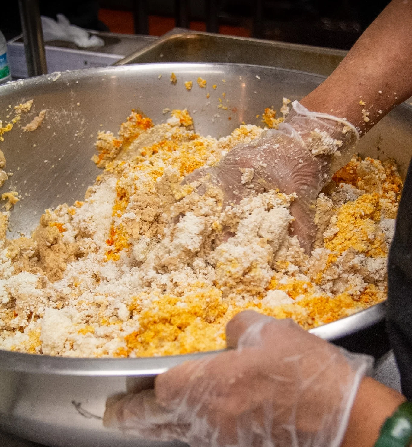 A large metal bowl filled with a mixed grain and color mixture, with a hand wearing a glove mixing the ingredients.
