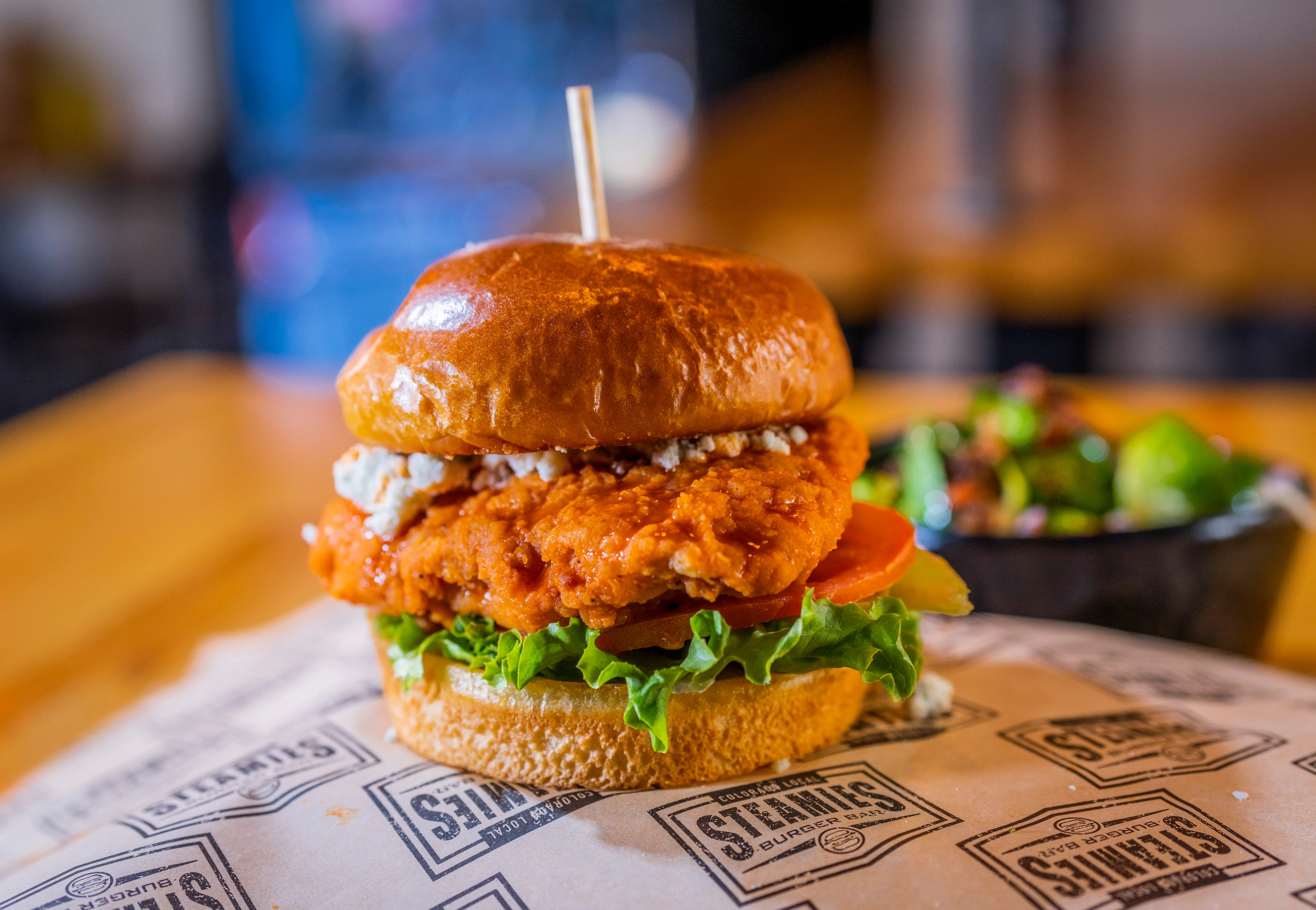 A crispy chicken sandwich with lettuce, tomato, and blue cheese crumbles on a toasted bun, with a side salad in the background.