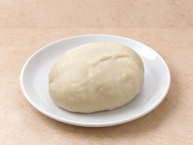 A round serving of fufu placed on a white plate. The fufu has a smooth, stretchy texture and is white in color.