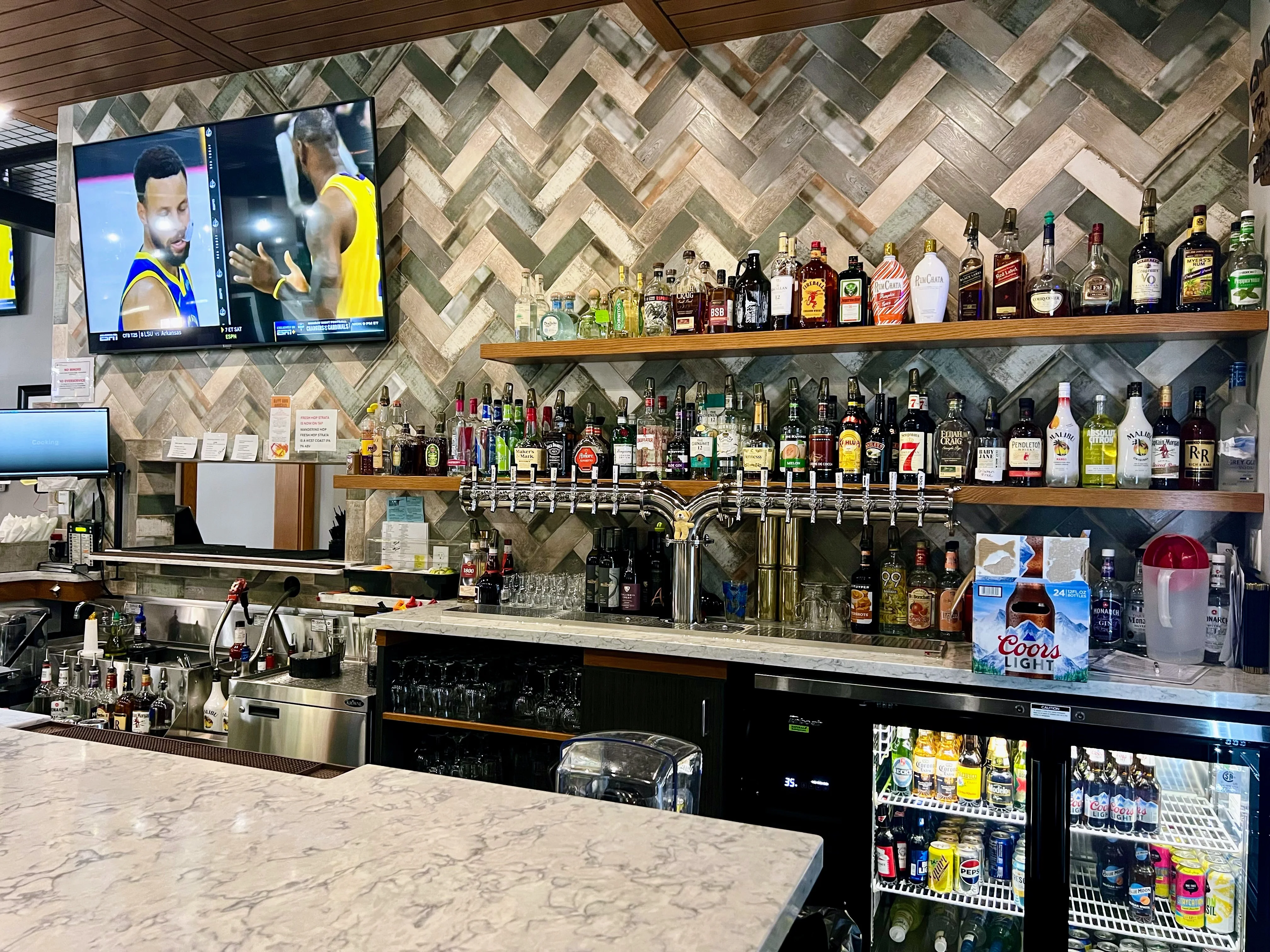 A well-stocked bar with various bottles of drinks on shelves, illuminated by soft lighting and featuring a large television above.