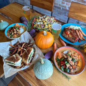 A vibrant assortment of food arranged on a wooden table, including sandwiches, colorful salads, and seasonal vegetables. The setting also includes two pints of beer and decorative pumpkins.