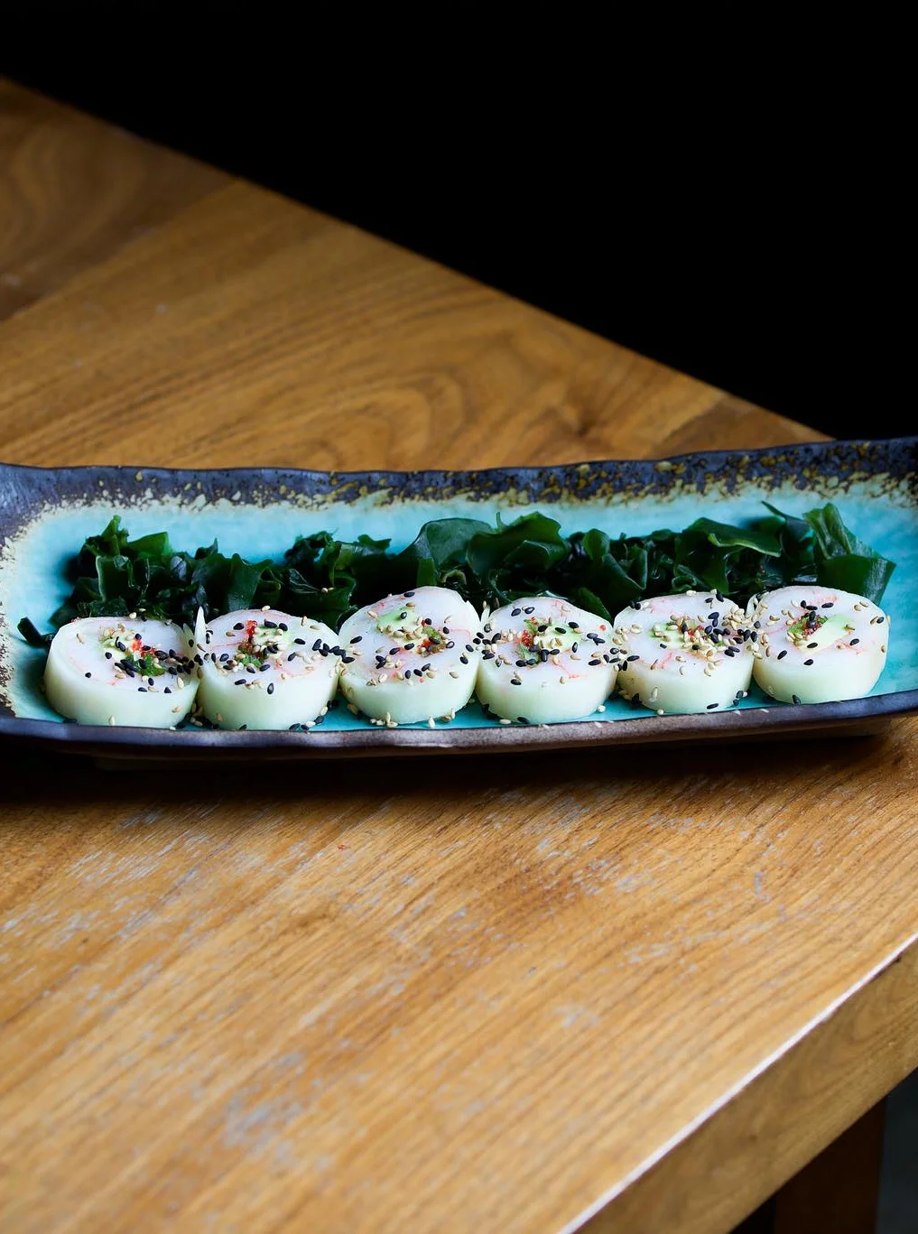 Cucumber-wrapped sushi rolls topped with sesame seeds and microgreens, served on a turquoise rectangular plate with a side of seaweed salad.