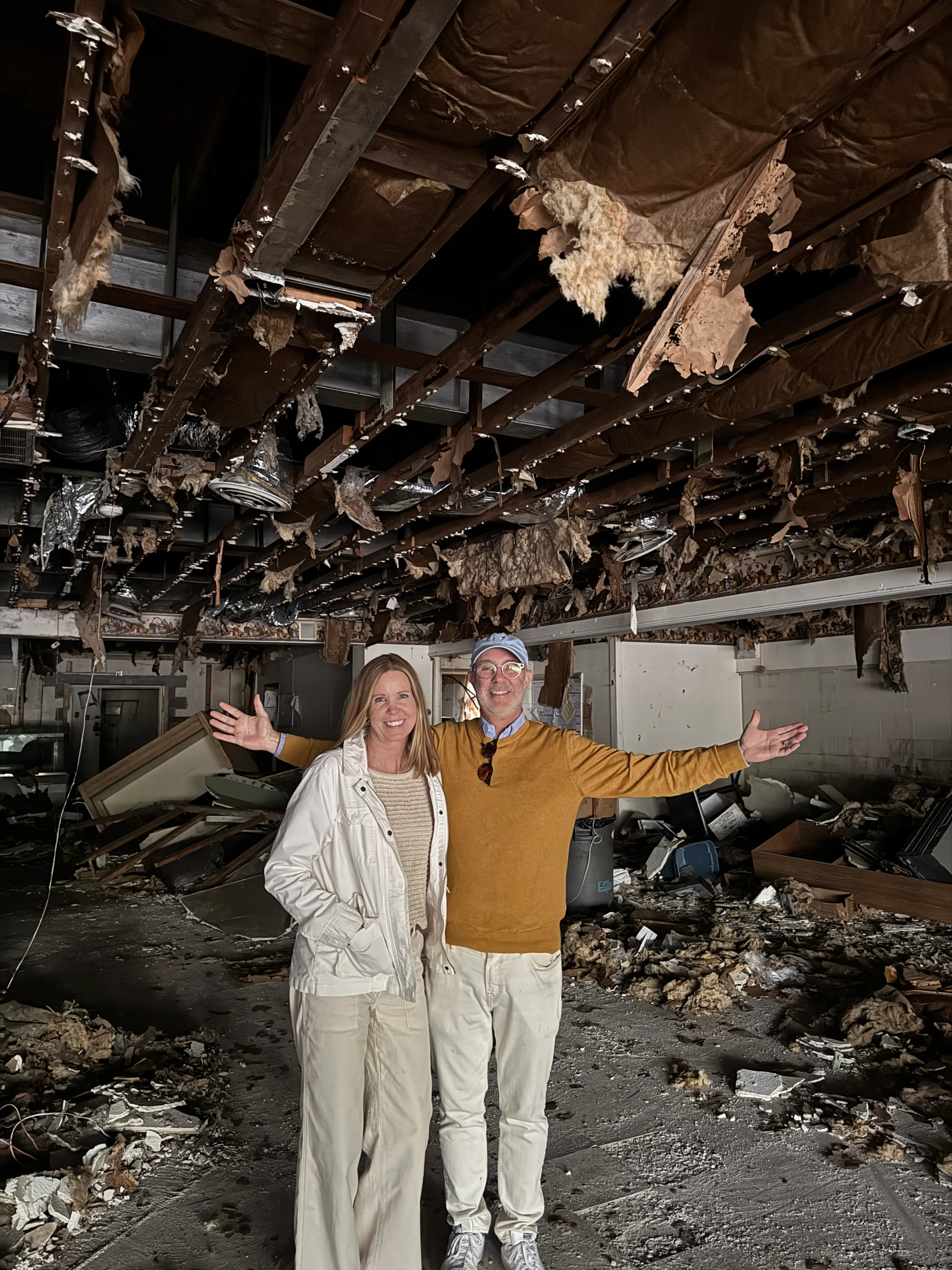 Two people smiling in a dilapidated interior space with debris.