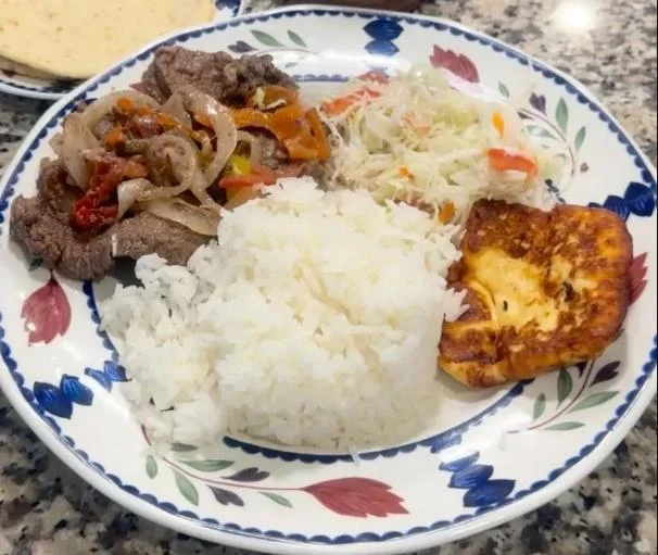 A vibrant meal featuring rice, seasoned beef with onions and peppers, coleslaw, and fried plantain on a decorative plate.