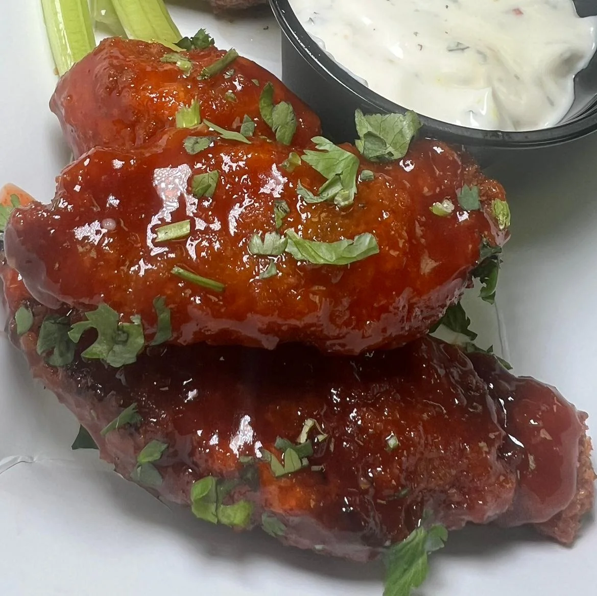 A plate of glazed chicken wings garnished with chopped herbs and served with a side of ranch dressing and celery sticks.