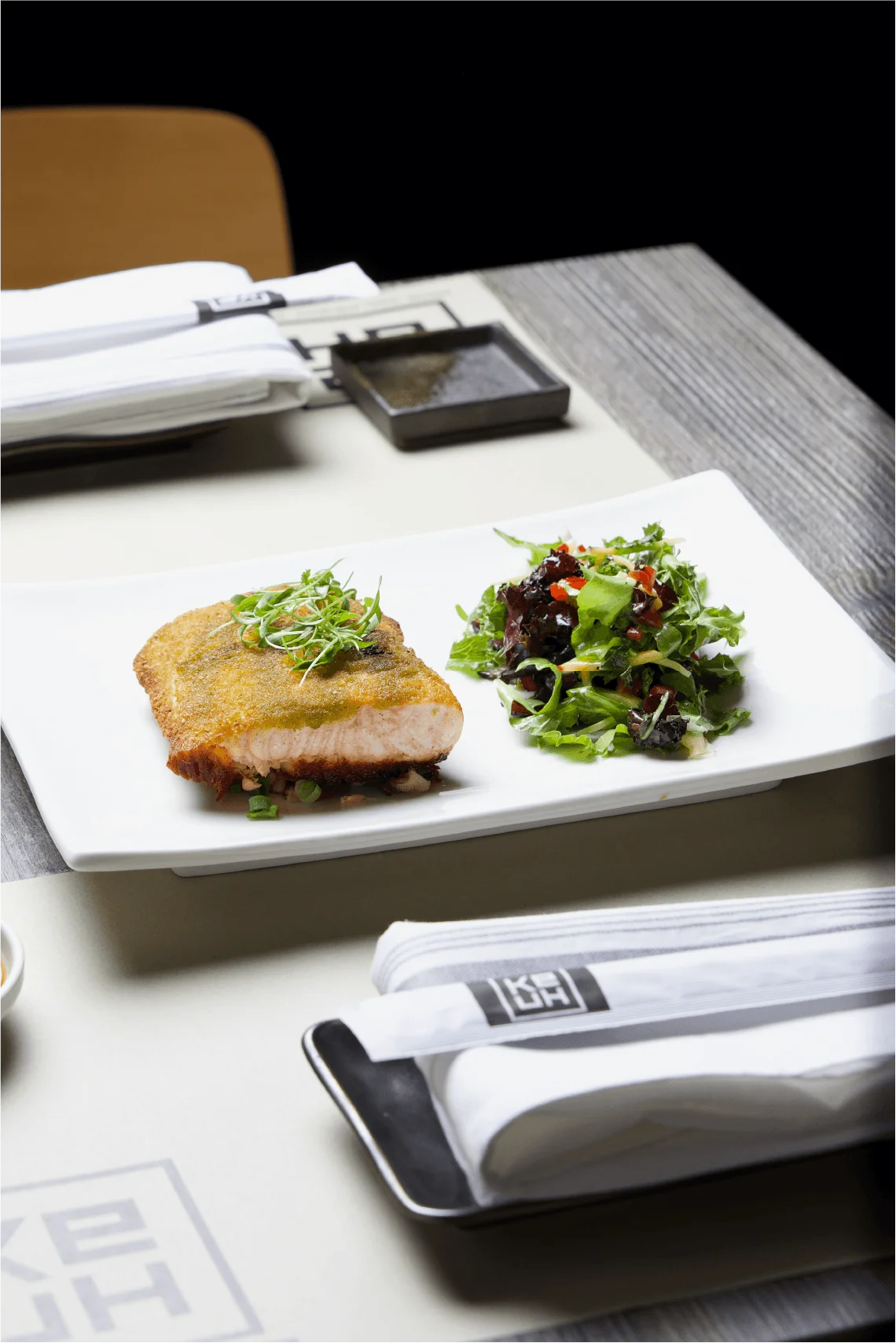 A crisp, herb-crusted salmon fillet garnished with microgreens and served alongside a vibrant mixed green salad on a rectangular white plate, set on a modern restaurant table with branded napkins.