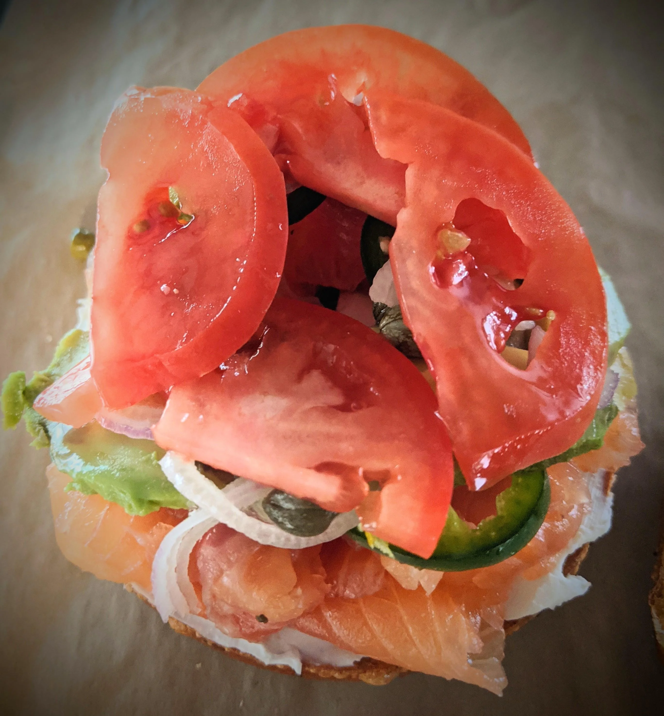 A sandwich topped with slices of fresh tomato and garnished with various fresh vegetables on a light background.