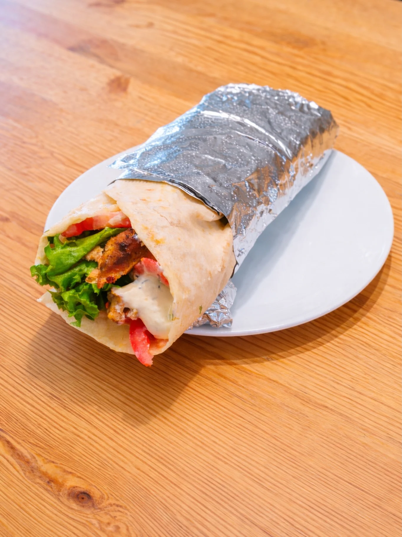 A wrapped sandwich with grilled chicken, lettuce, and tomato, partially revealed from its foil wrapping and set on a white plate.