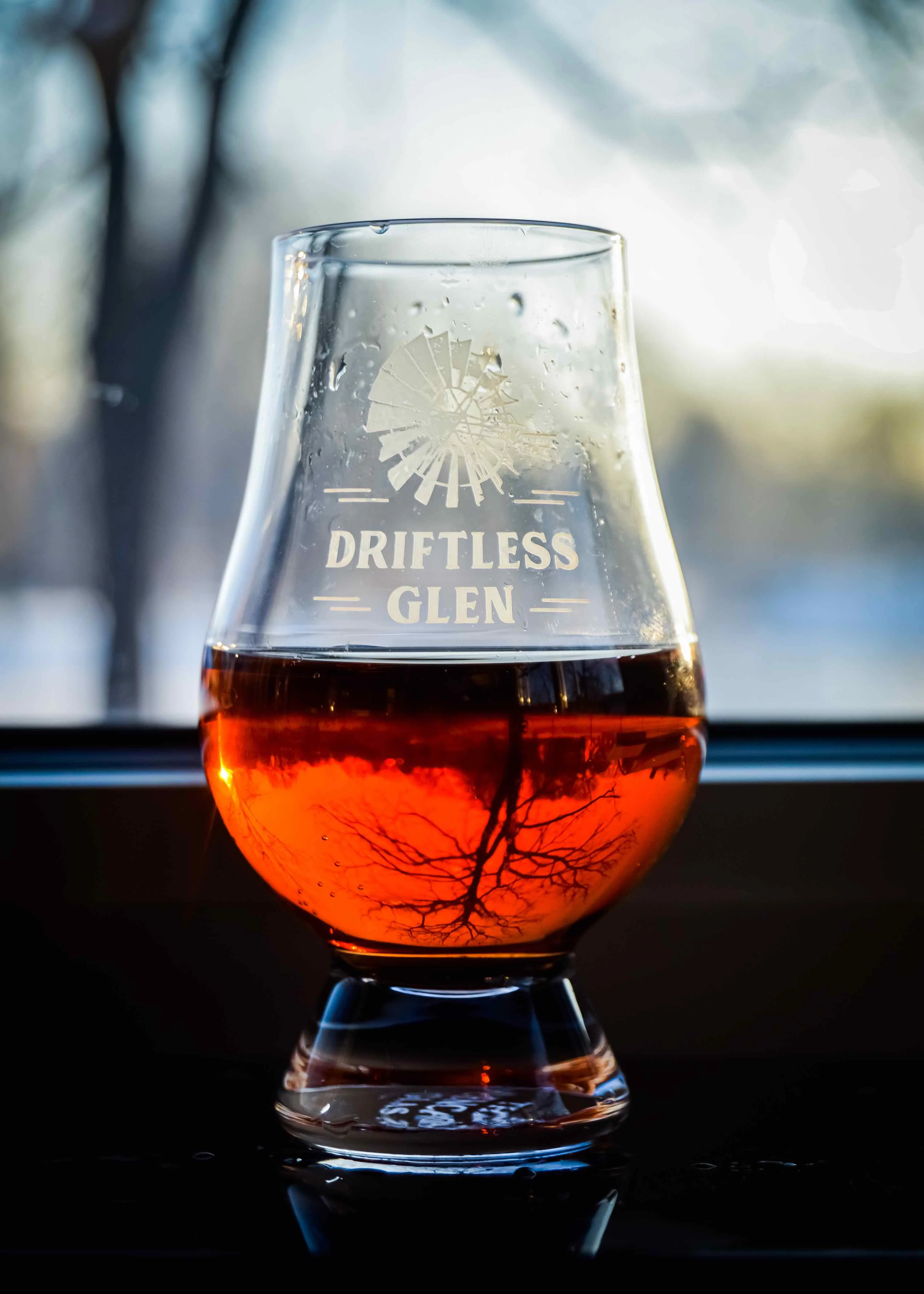 A Glencairn full of whiskey sits in front of a window with the reflection of branches distorted through the reflection