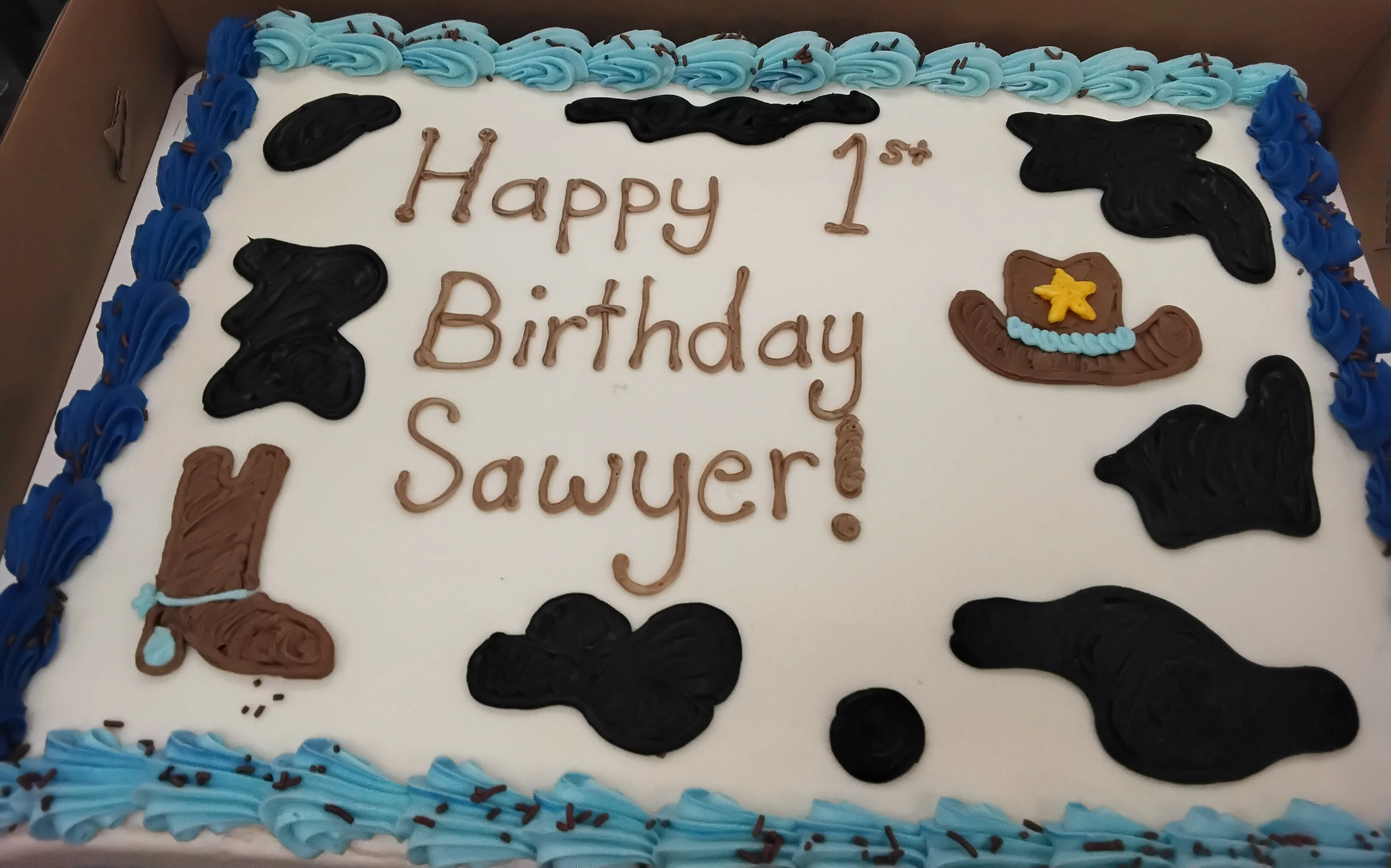 Sheet cake with light & dark blue borders, black cow print, a cowboy hat, & boot drawn in icing that says "Happy 1st birthday Sawyer!"