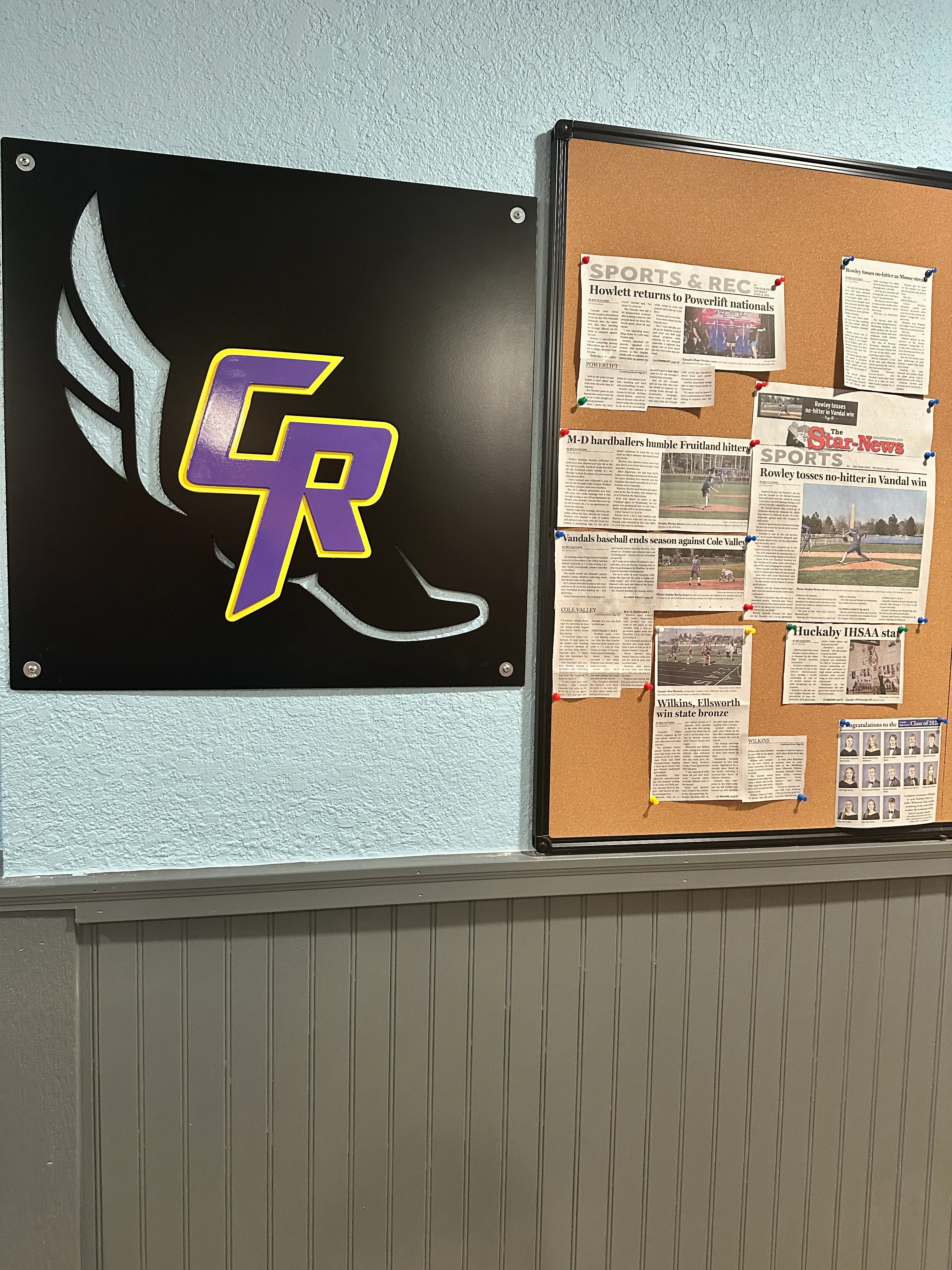 A wall display featuring a logo with wings and the initials 'CR' in purple and yellow, alongside a bulletin board filled with newspaper clippings about sports events.