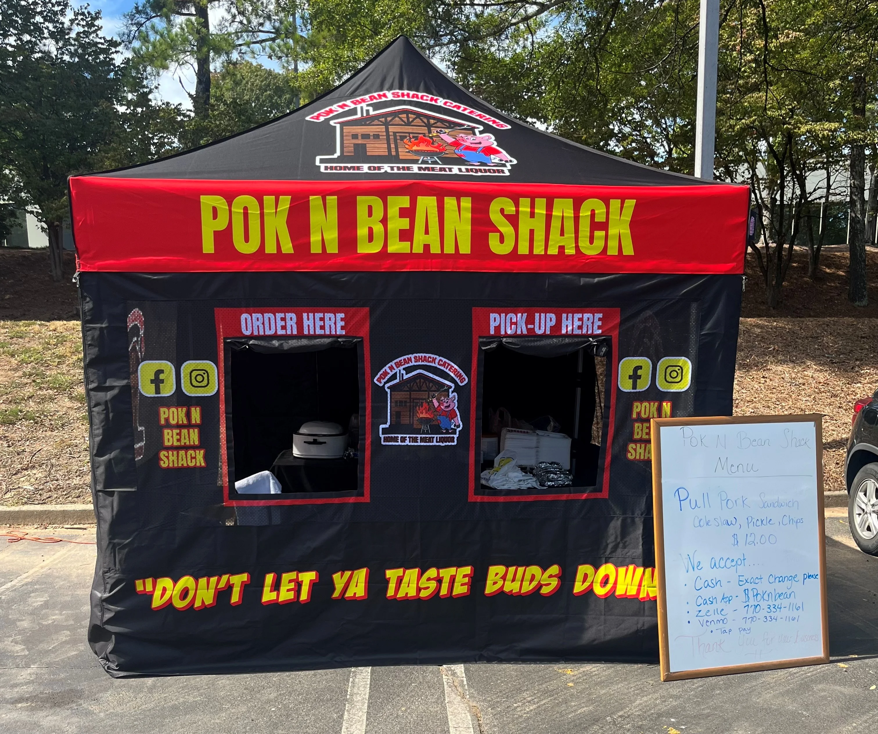 A food shack with a vibrant red and black awning, advertising 'Pok N Bean Shack' and featuring a menu for pulled pork sandwiches. A whiteboard lists payment options and prices.