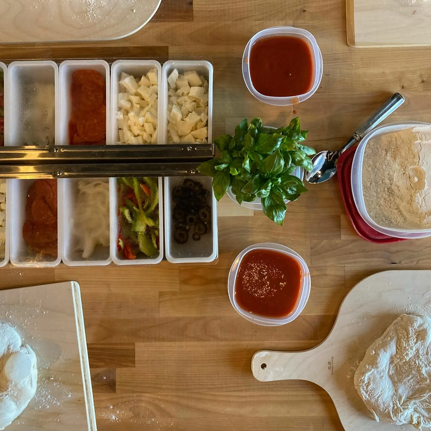 Various ingredients for pizza preparation displayed on a wooden surface, including sauces, vegetables, and cheese.