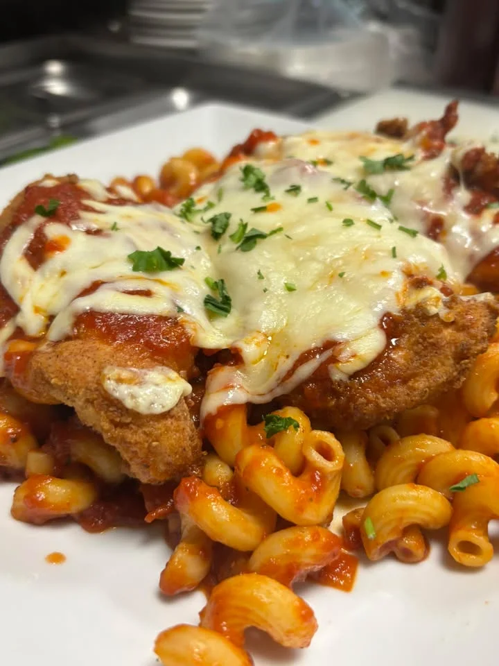 A plate featuring chicken Parmesan served over cavatappi pasta with marinara sauce and melted cheese, garnished with parsley.