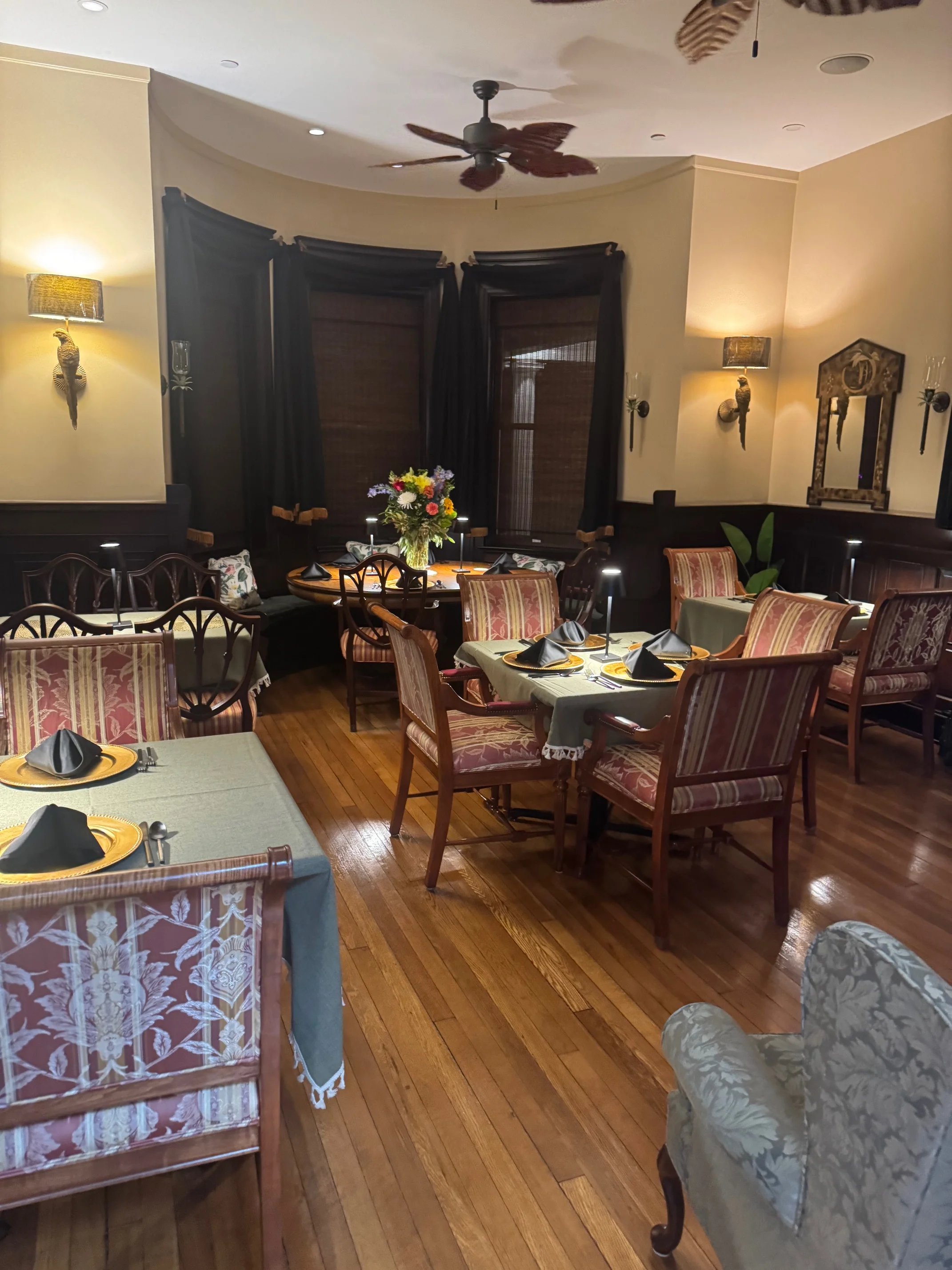 A dining area with tables set elegantly, featuring decorative tableware and a floral centerpiece. Natural light filters through the stylish window treatments.