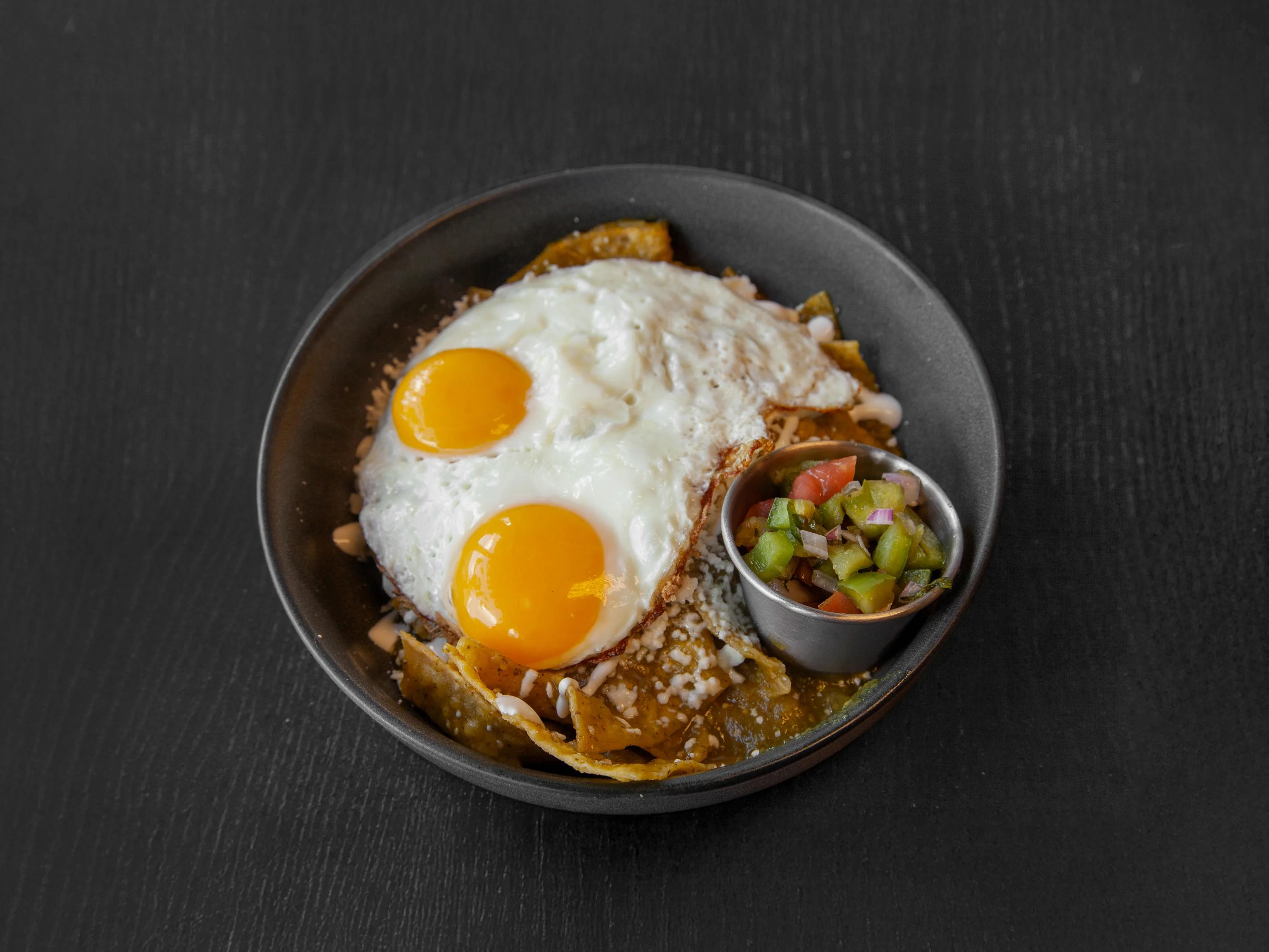 A bowl of chilaquiles topped with two sunny-side-up eggs and a side of salsa, served with crispy tortilla chips.