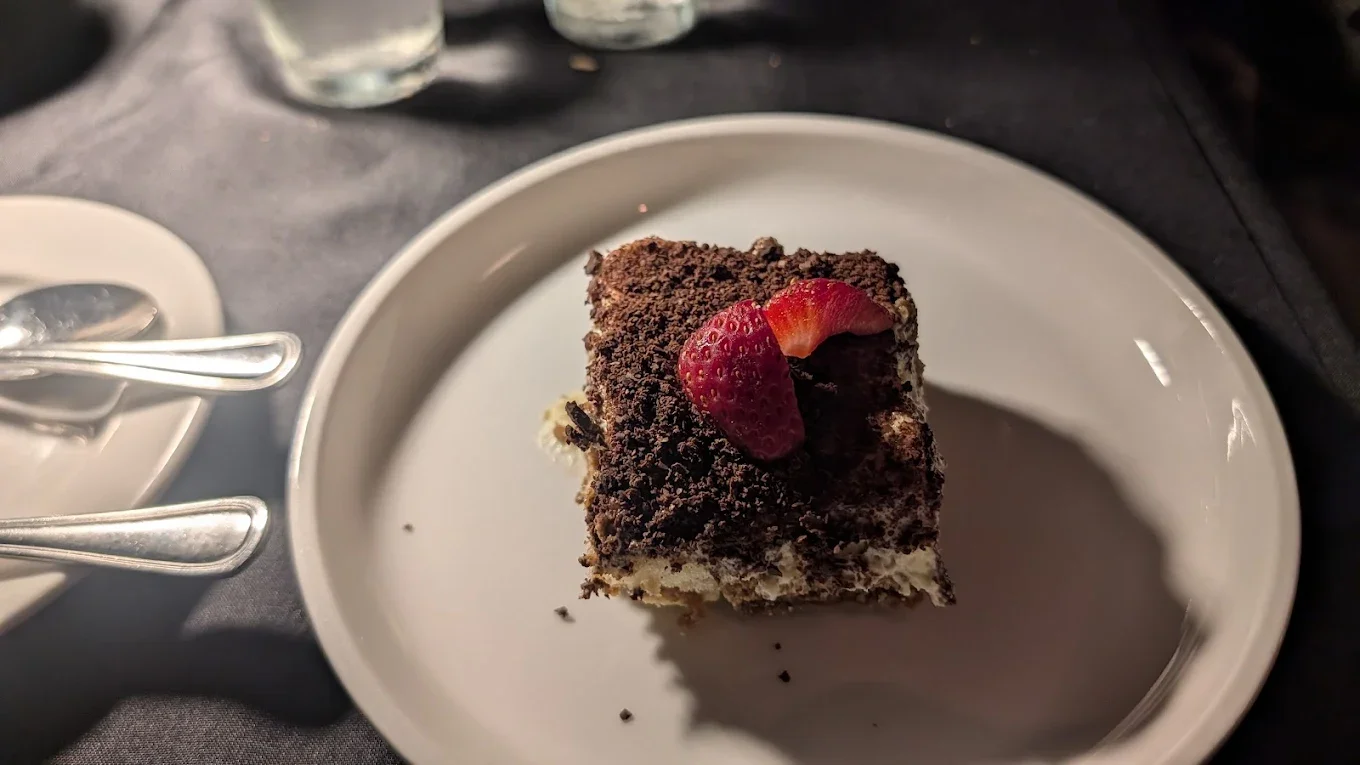A slice of chocolate dessert topped with a strawberry on a white plate, resting on a dark table.