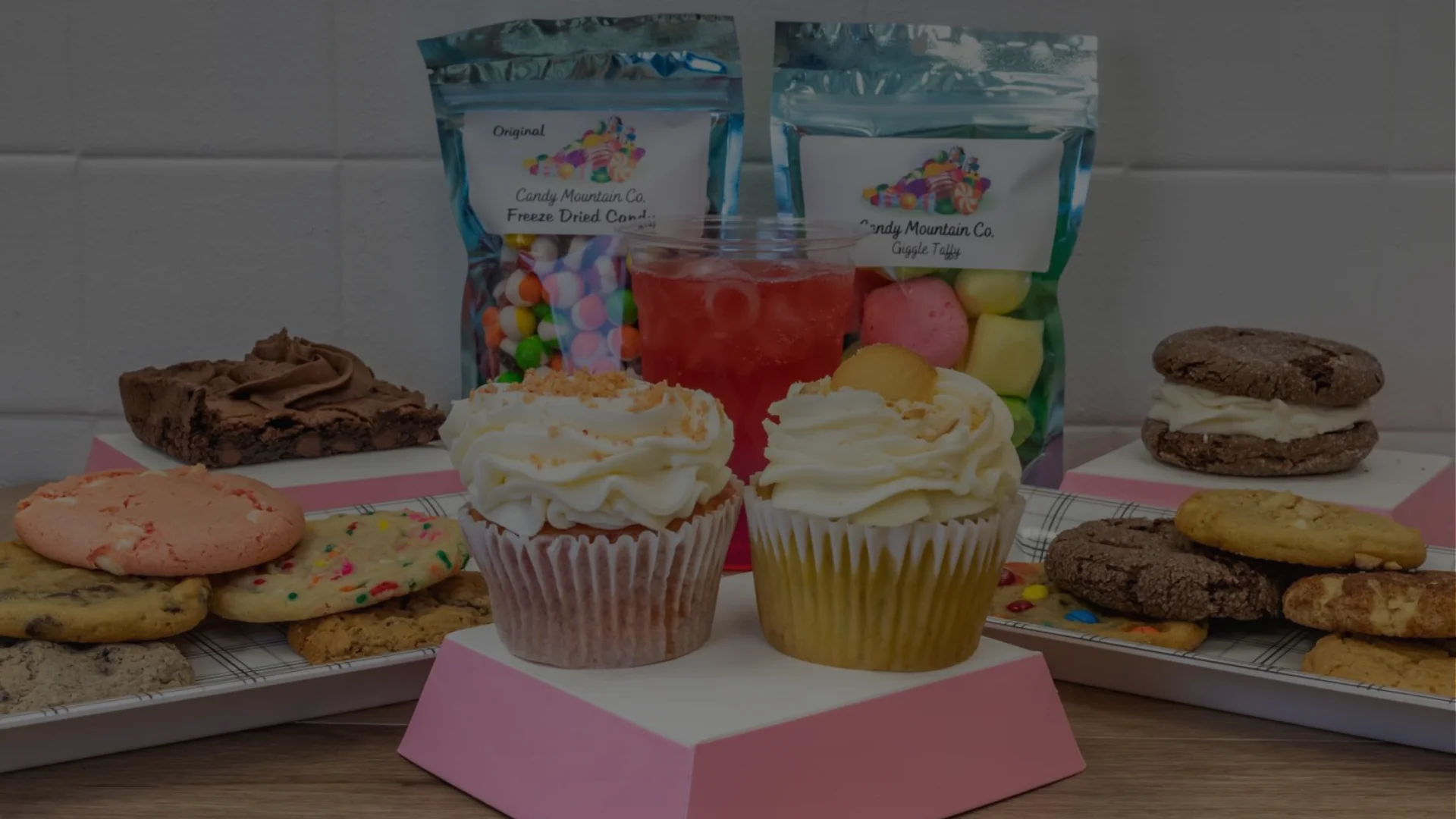 A dessert spread with cupcakes, cookies, brownies, and packaged candy, displayed with drinks on a table.