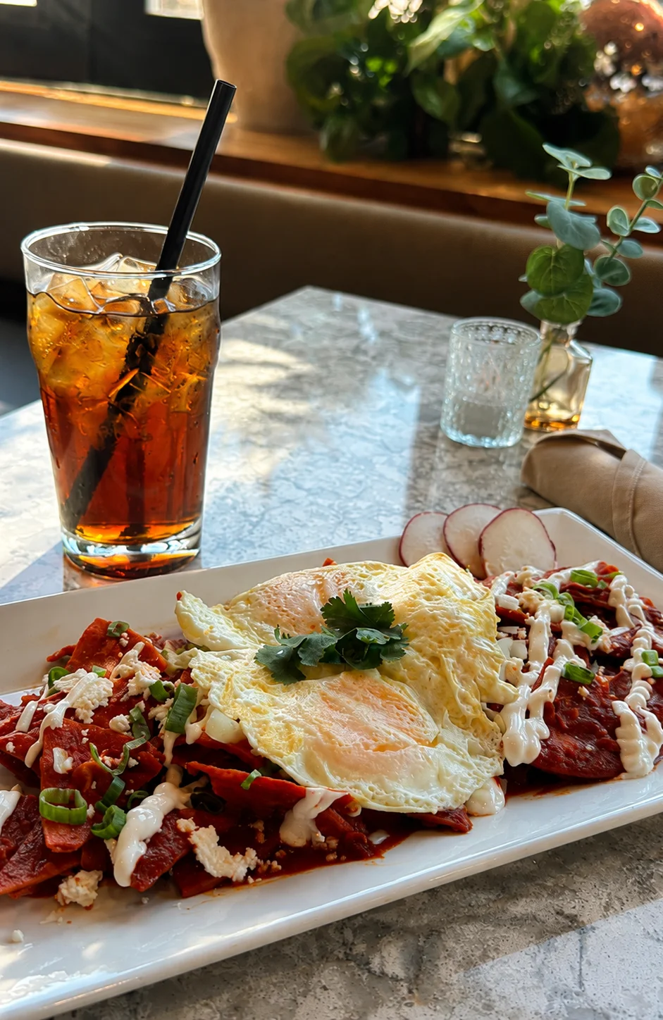 A plate of tacos filled with seasoned meat, topped with diced onions, cilantro, and fresh mango, served with lime wedges and a variety of sauces. A drink with ice is visible in the background.