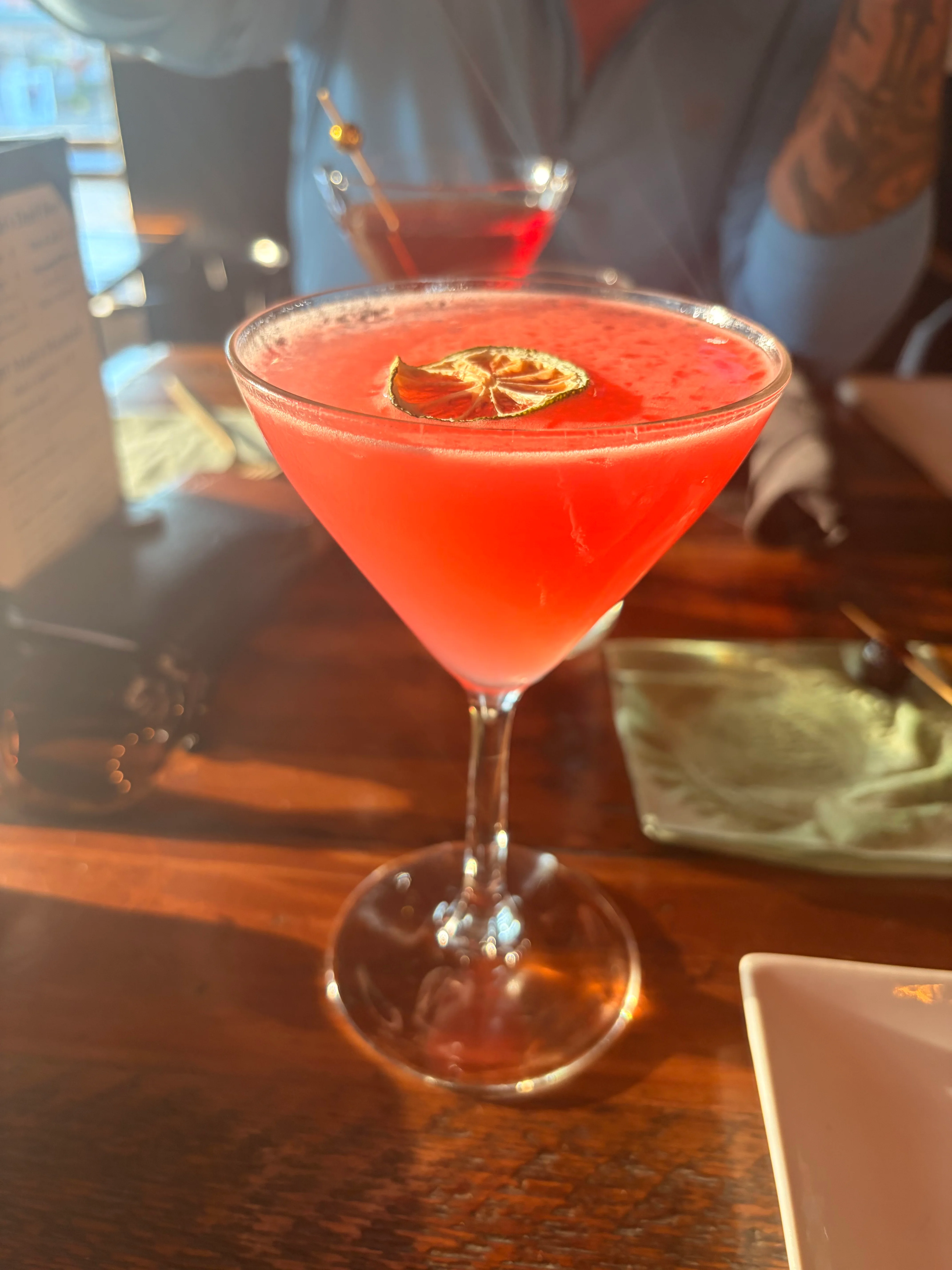 A cocktail in a coupe glass, garnished with a pine cone, with a blurred background of flowers.