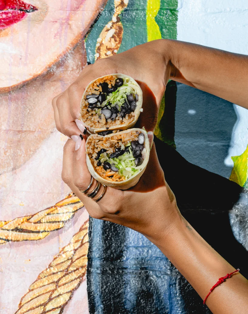 Hands holding a burrito cut in half, filled with rice, black beans, and lettuce in front of a Frida Kahlo mural at Friducha in Brooklyn, NY.