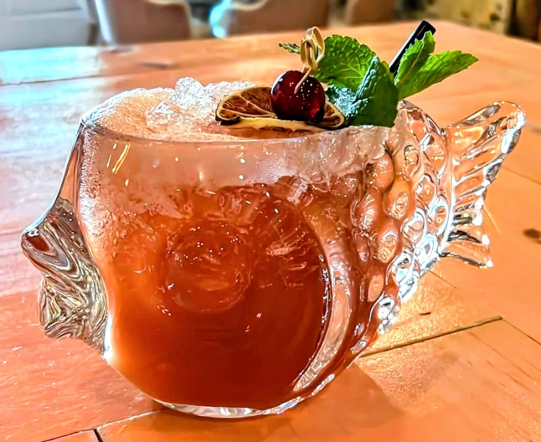 A colorful cocktail served in a fish-shaped glass with mint and cherry garnish.