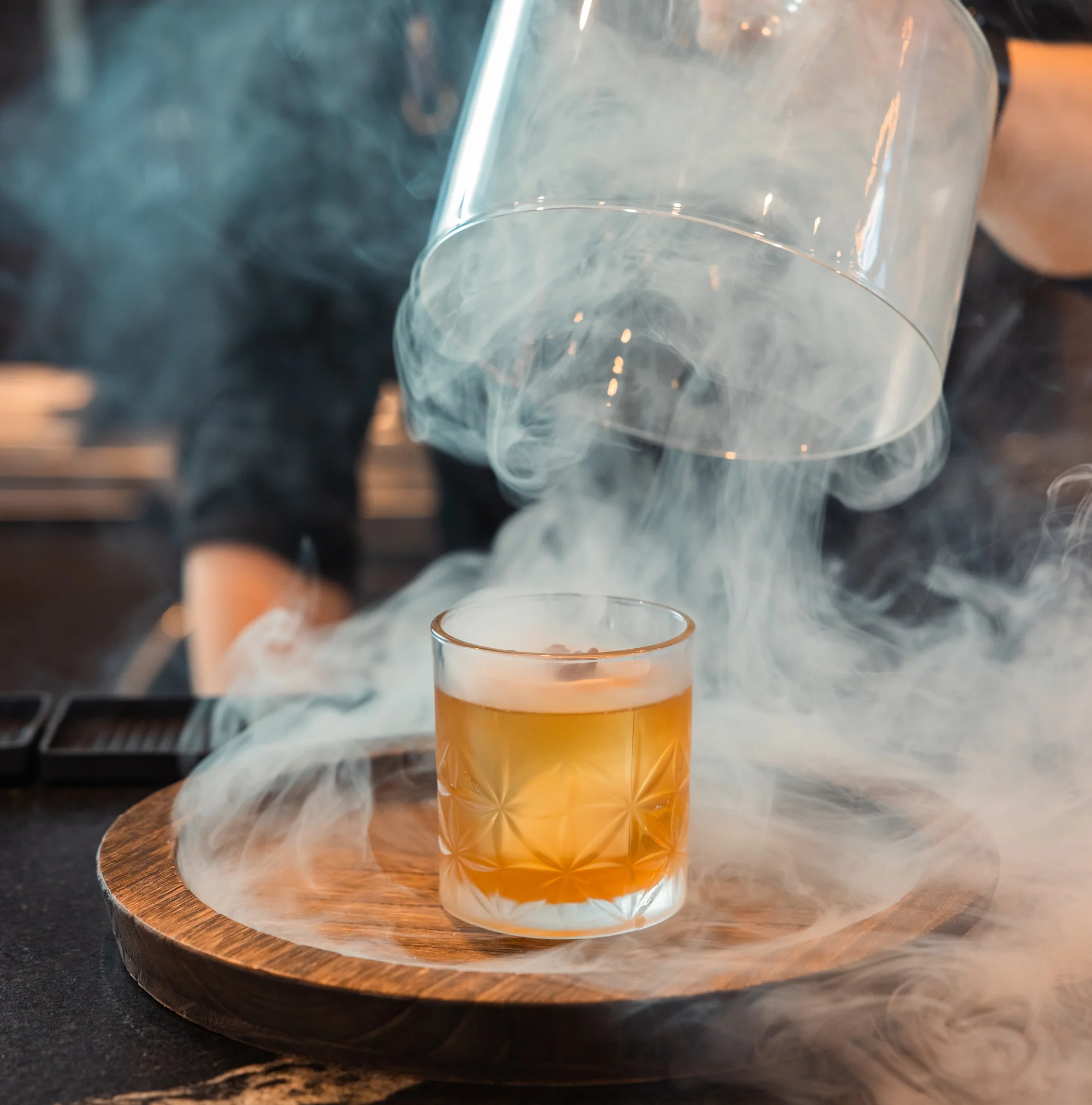 Bartender pouring smoke over a cocktail in a glass on a wooden tray.