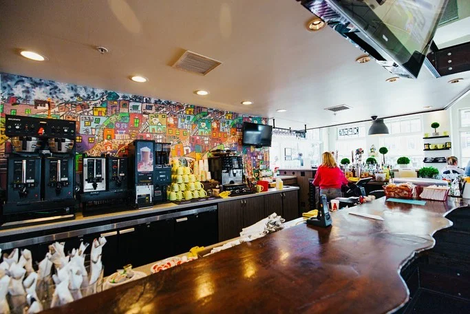 A coffee preparation area featuring multiple machines and a bright atmosphere. A lone customer is seen near the counter, with a colorful mural in the background.