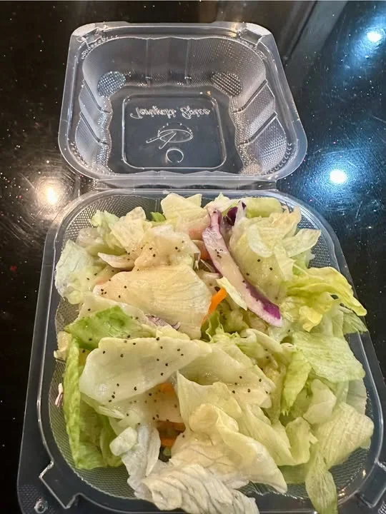 A clear plastic container filled with mixed salad greens, accompanied by some shredded vegetables and dressed with black pepper.