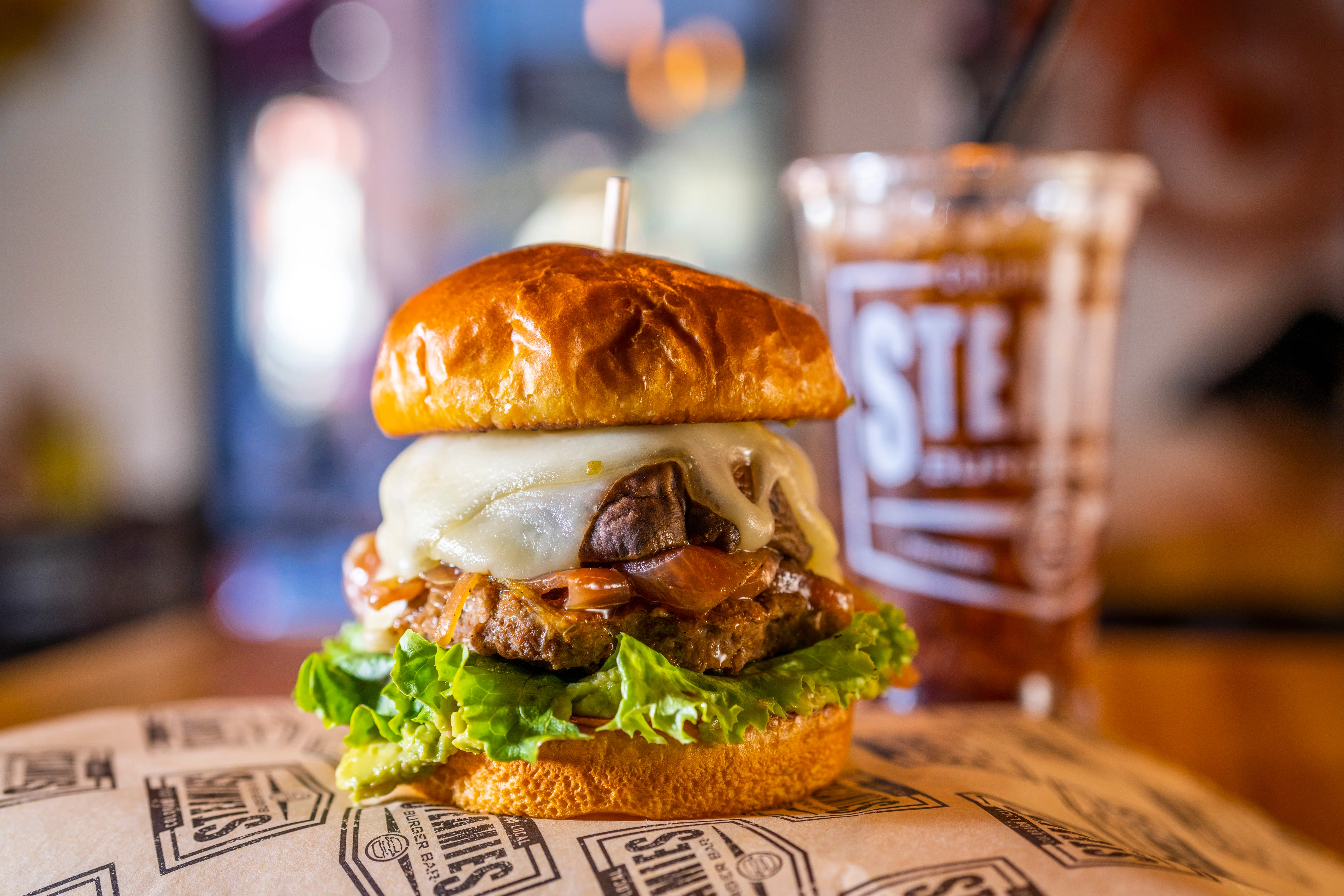 A close-up of a gourmet burger featuring a toasted bun, lettuce, cheese, and grilled meat, accompanied by a drink in the background.