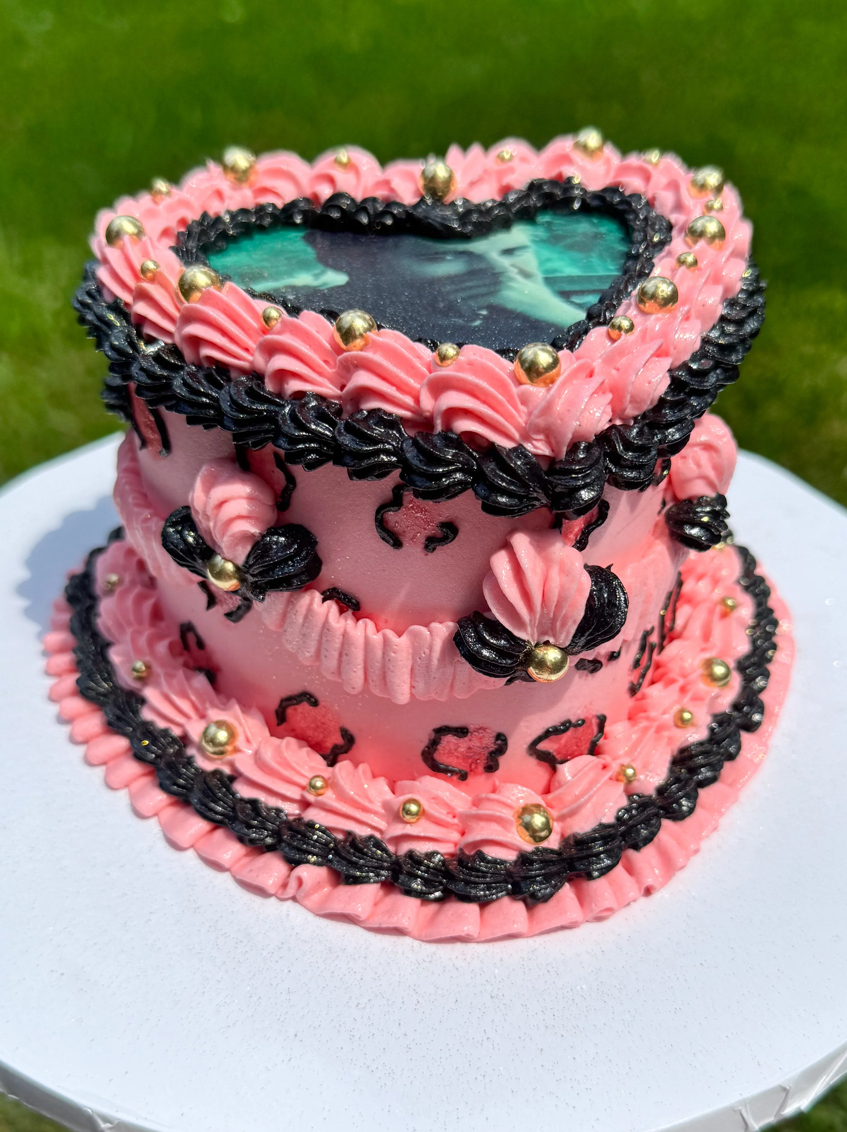 A heart-shaped cake decorated in pink and black frosting, featuring gold accents and a photo on top.