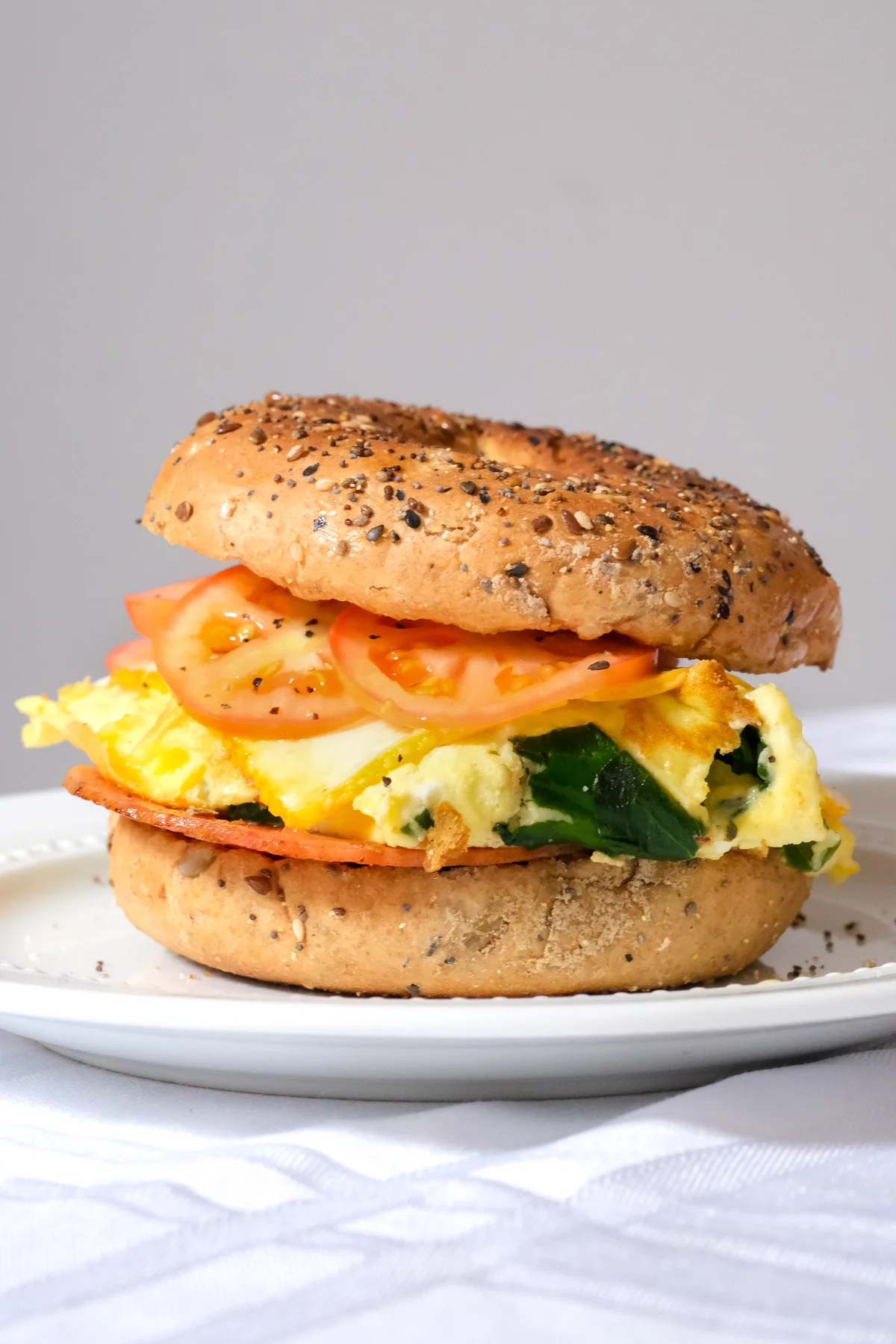 A bagel sandwich filled with scrambled eggs, spinach, and tomato slices, topped with poppy seeds.