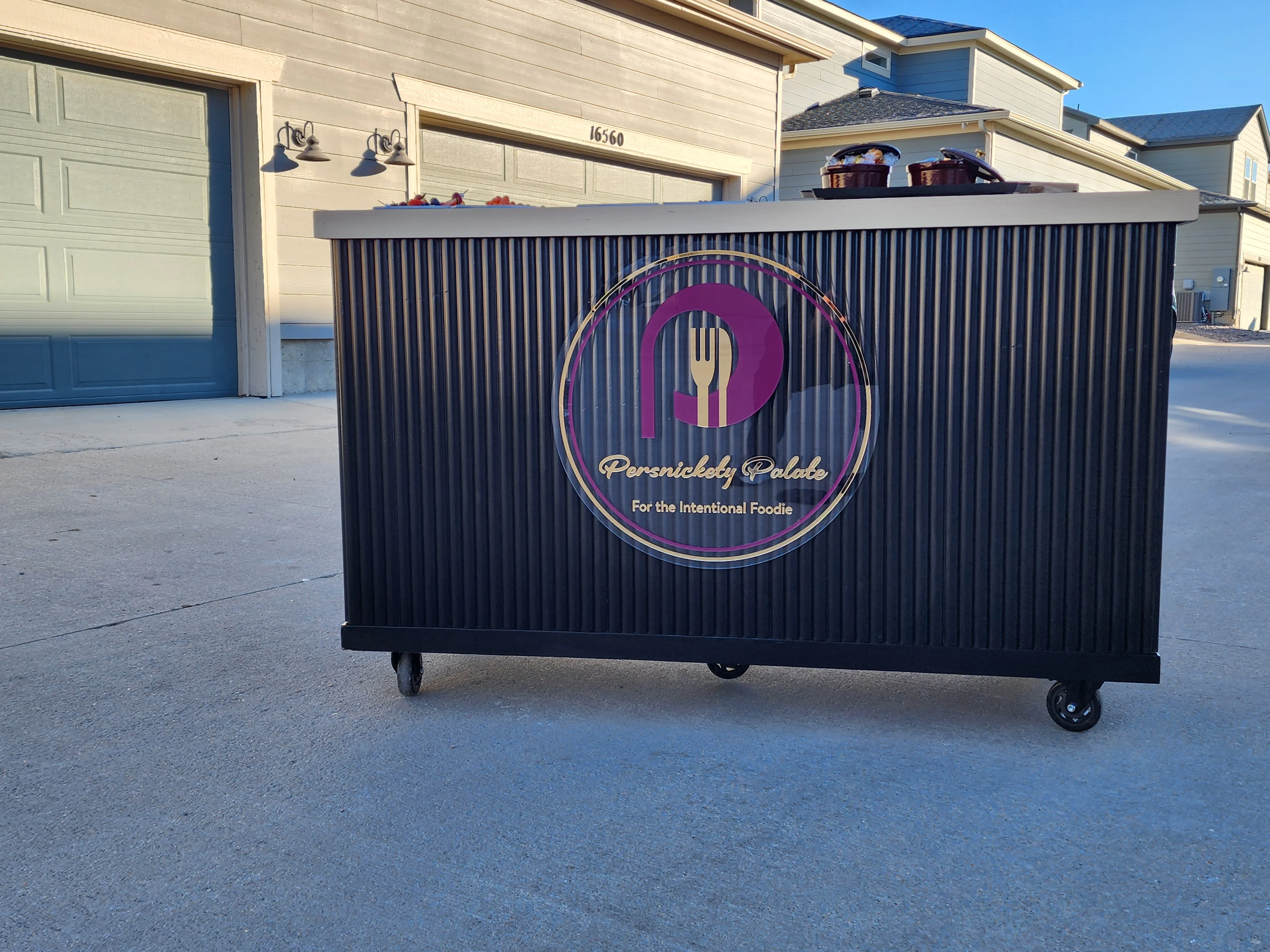 A black food cart with a logo and topped with small cooking pots. The cart is situated in a residential area.