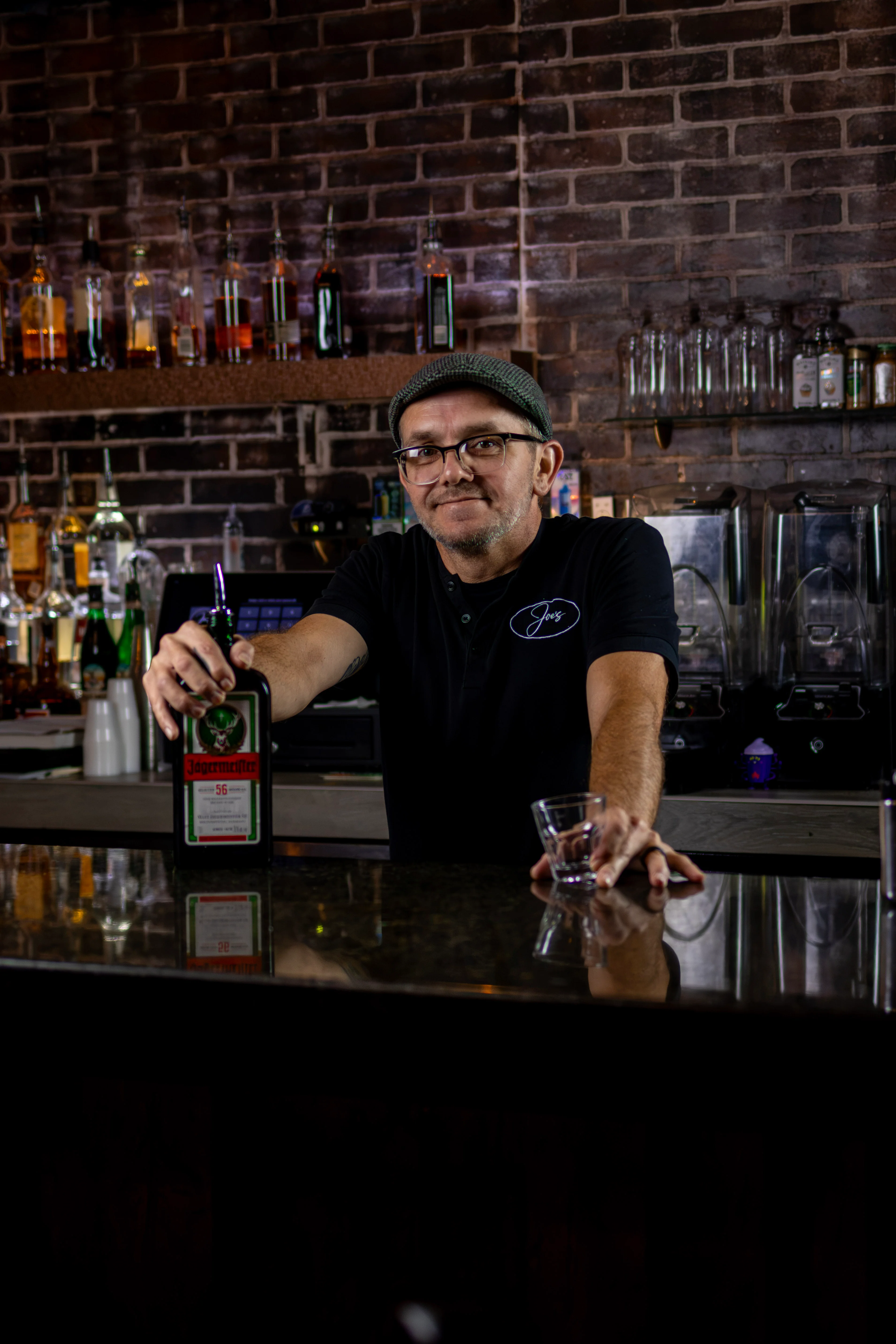 Bartender behind a bar with liquor bottles and exposed brick wall