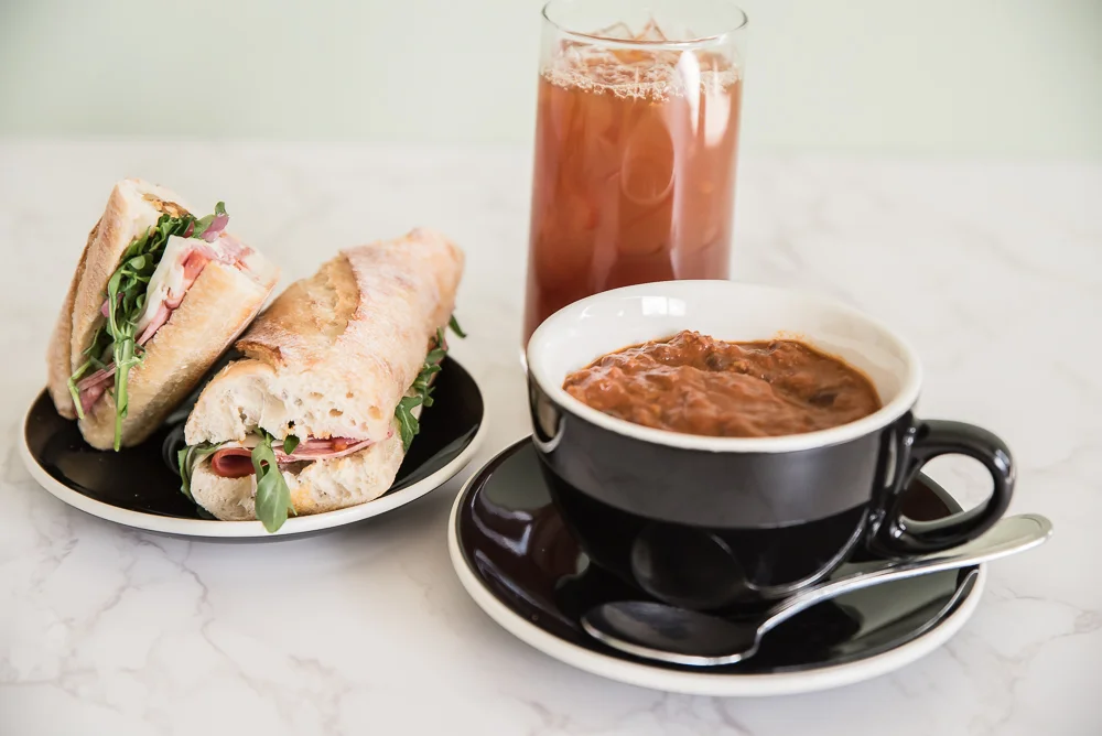 A fresh baguette sandwich with meat and greens sits on a plate beside a glass of iced tea and coffee, creating an inviting and satisfying meal.