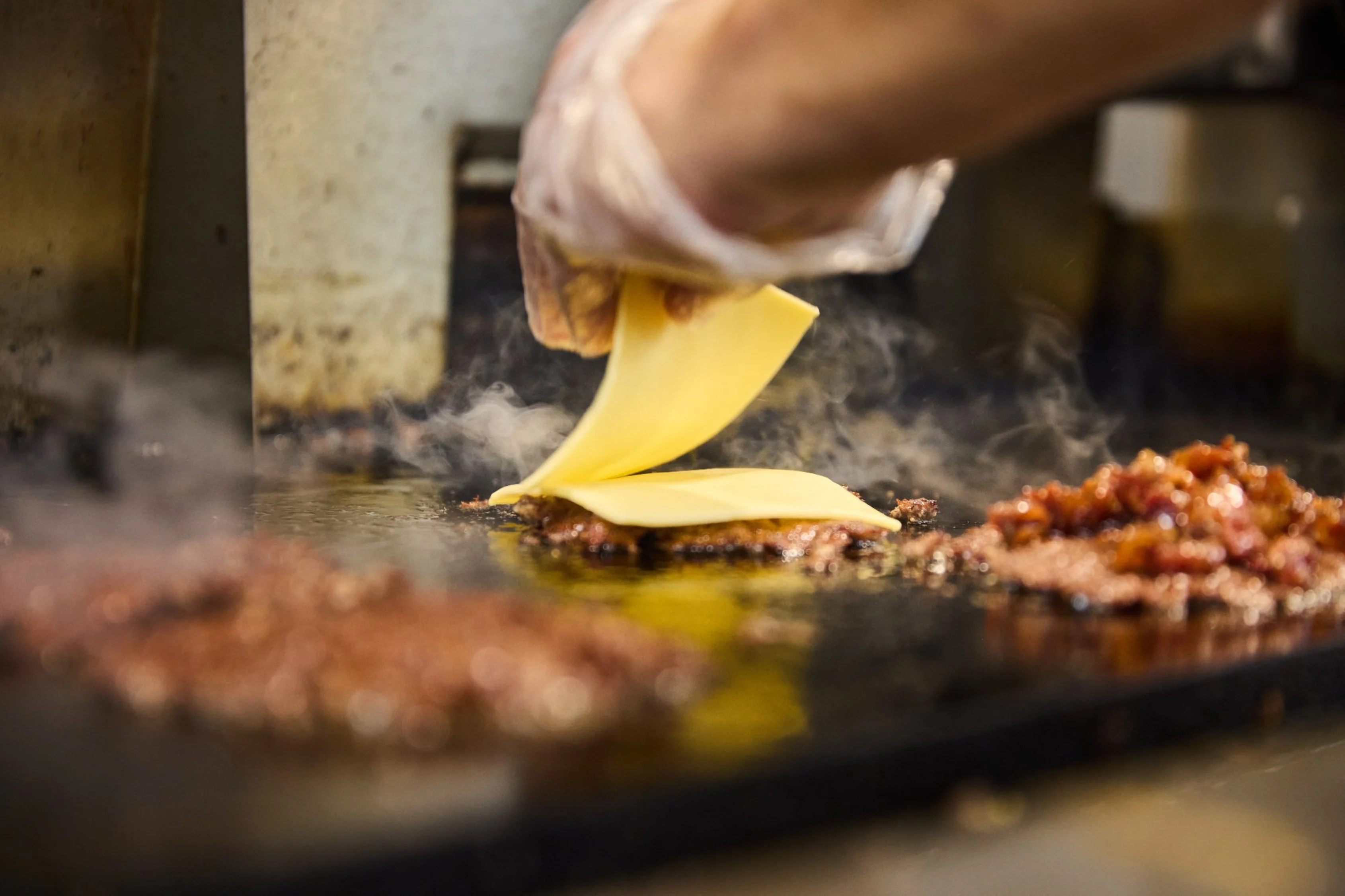 A hand placing a slice of cheese on top of sizzling meat on a grill, with steam rising around it.