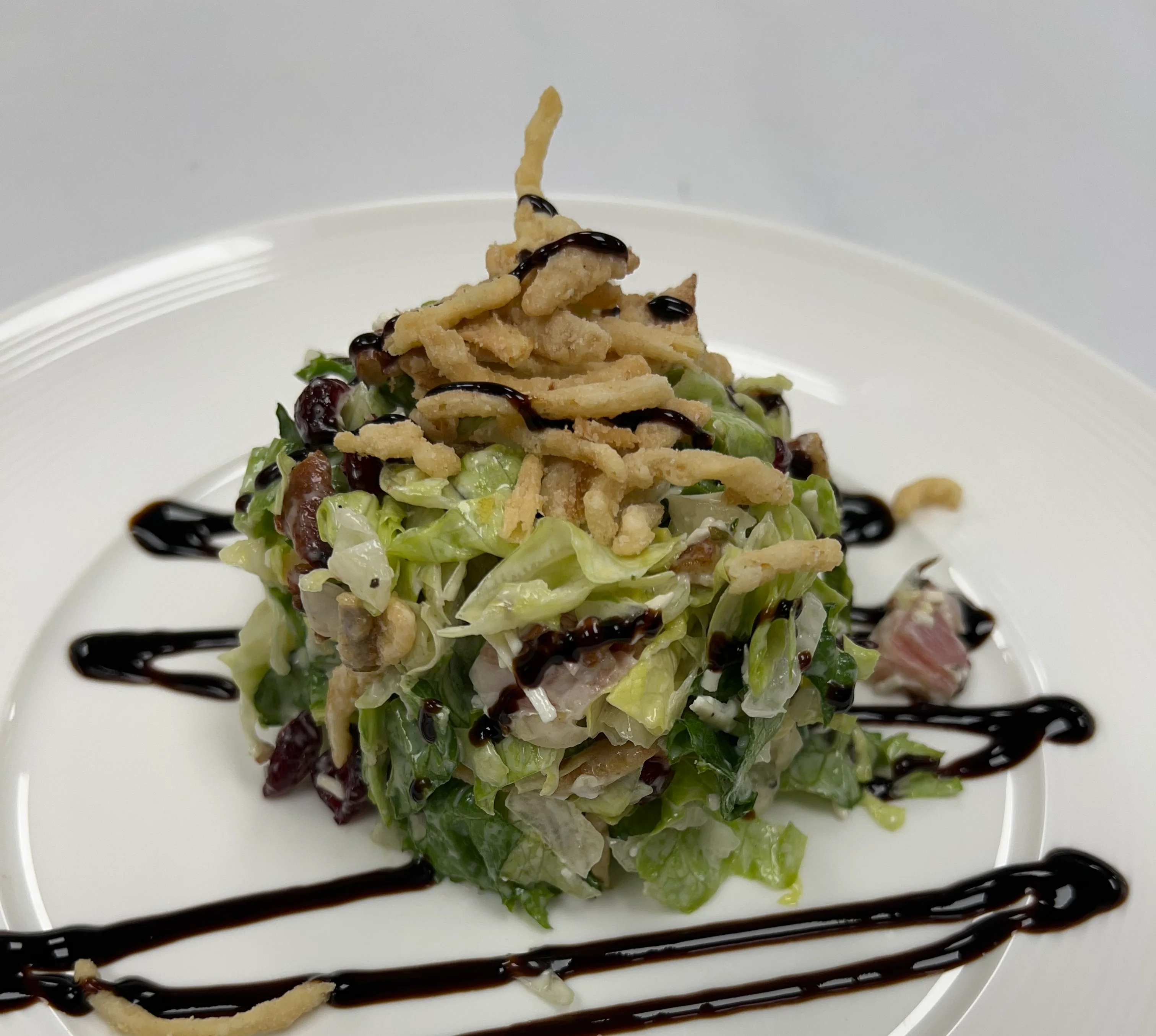 A neatly stacked chopped salad made with crisp romaine lettuce, dried cranberries, and bits of bacon, topped with crunchy fried onions and a drizzle of dark balsamic glaze. The salad is plated on a white dish with artistic lines of balsamic glaze and a few scattered fried onion pieces for garnish.