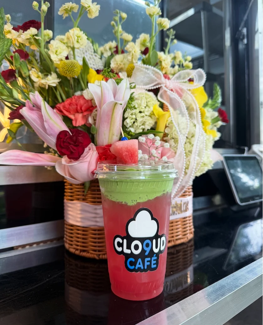 A vibrant drink with layers of red and green, topped with a piece of watermelon, placed beside a bouquet of colorful flowers.