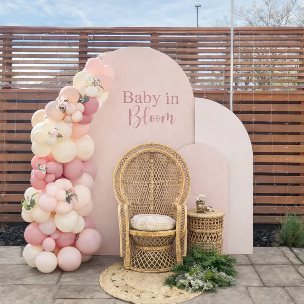 A decorative display featuring a rattan chair, a small side table, and a vibrant arrangement of balloons in shades of pink. The backdrop has the text 'Baby in Bloom'.