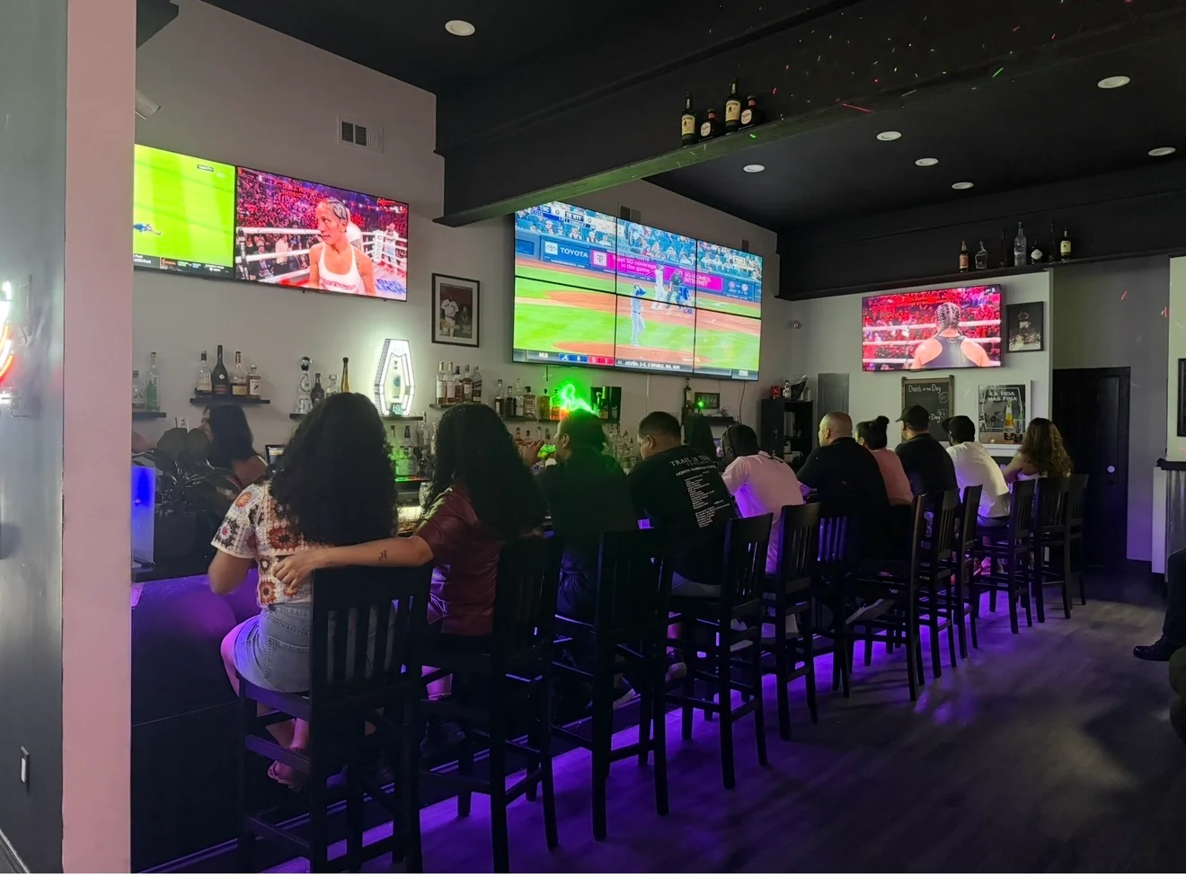 A lively bar scene with patrons seated at a long counter, multiple screens showing sports events, and a sleek interior design with colorful lighting.