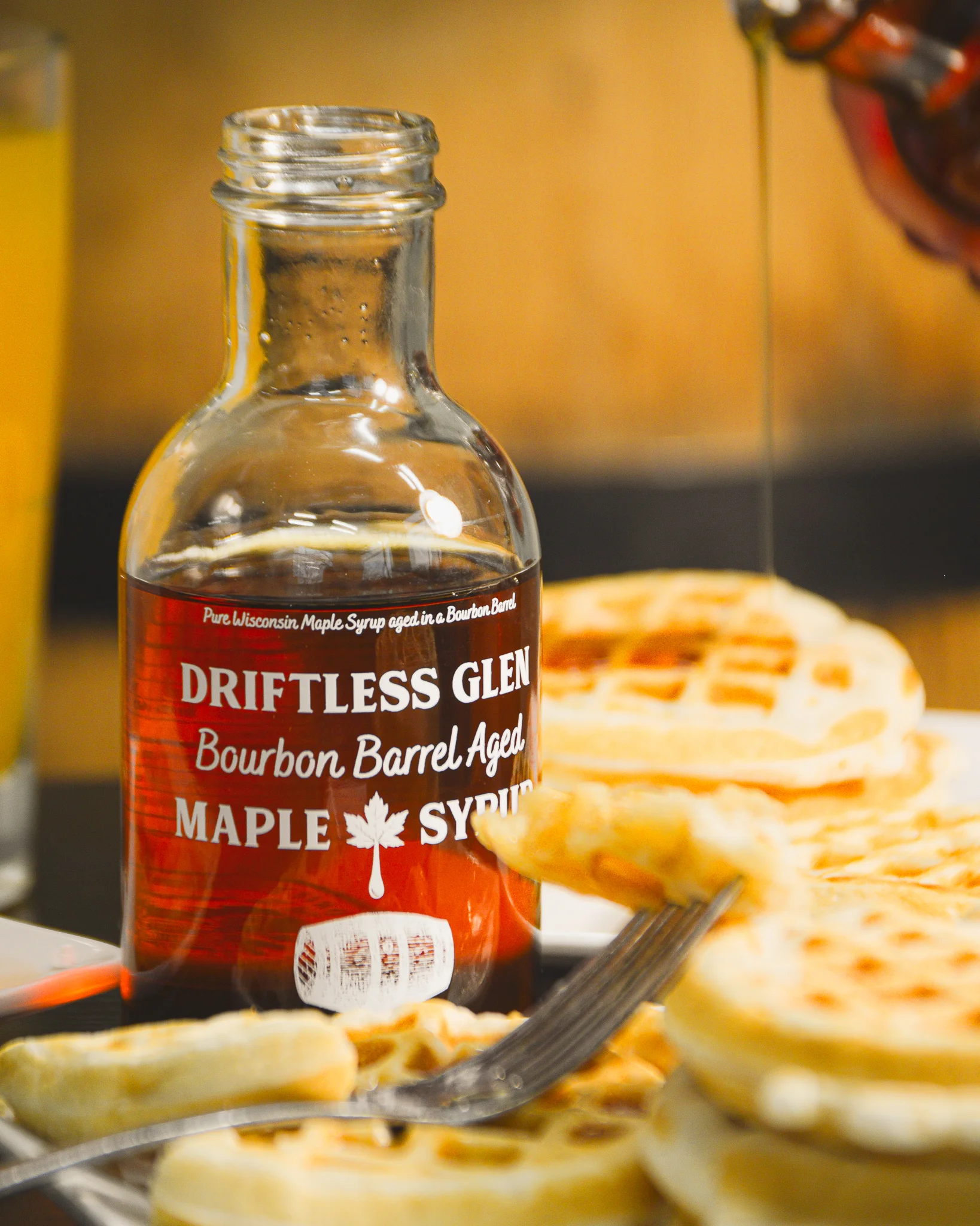 A bottle of maple syrup is surrounded by pancakes and a fork and syrup drizzled everywhere