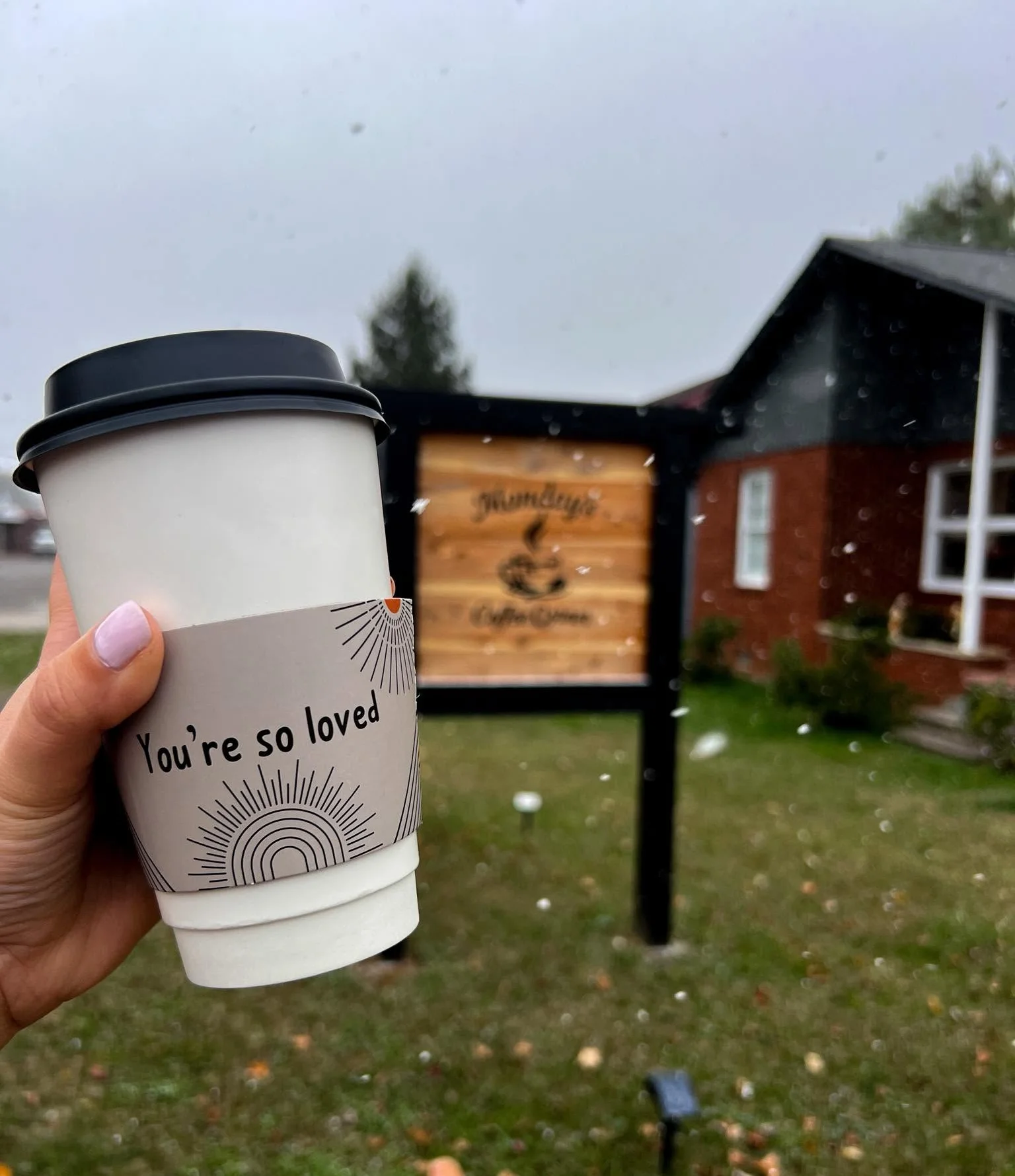 A hand holds a glass of coffee with leaf-shaped latte art in front of green leaves.