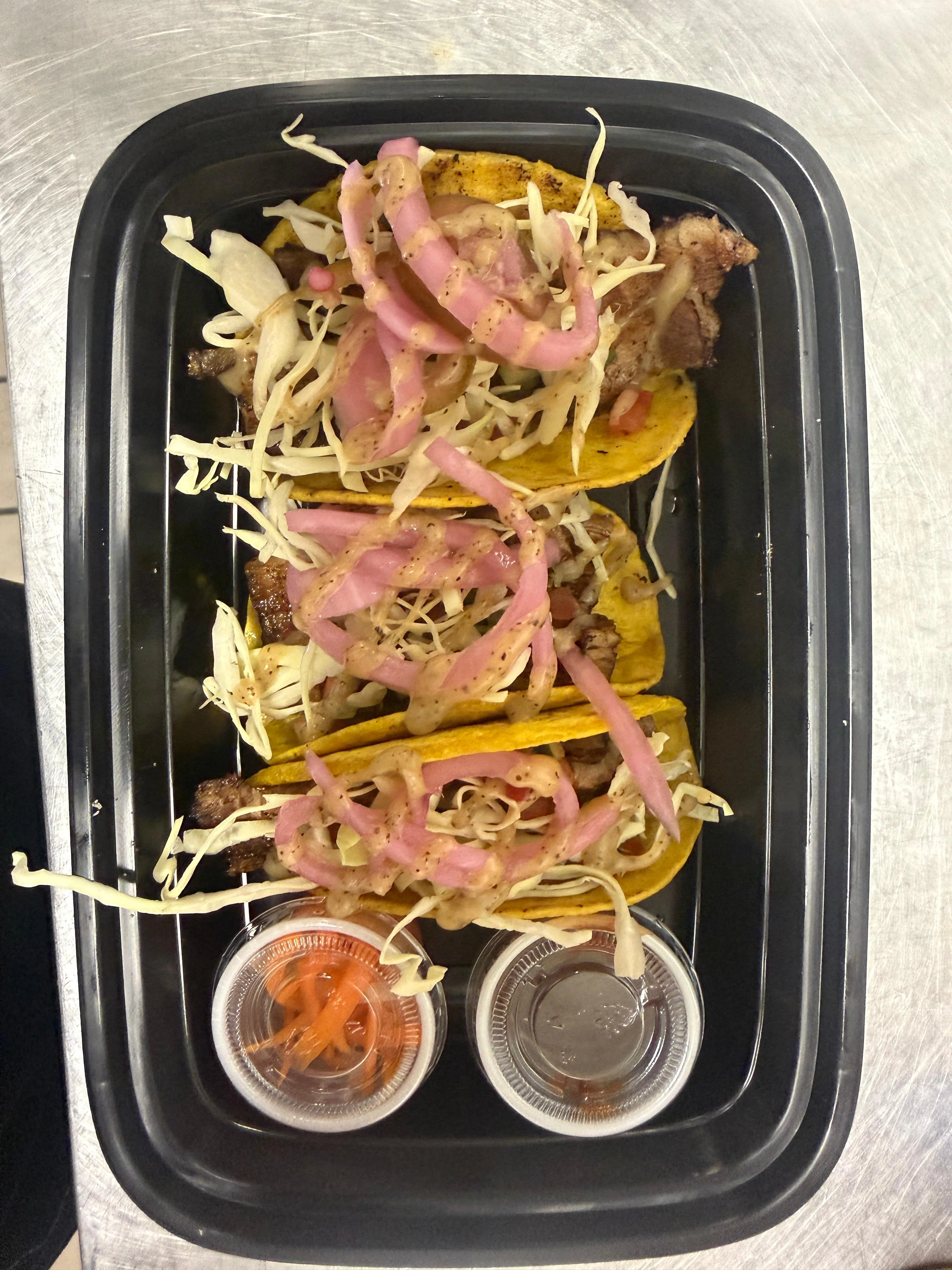 Three tacos with various toppings including shredded cabbage, pickled onions, and served with two small sauces.
